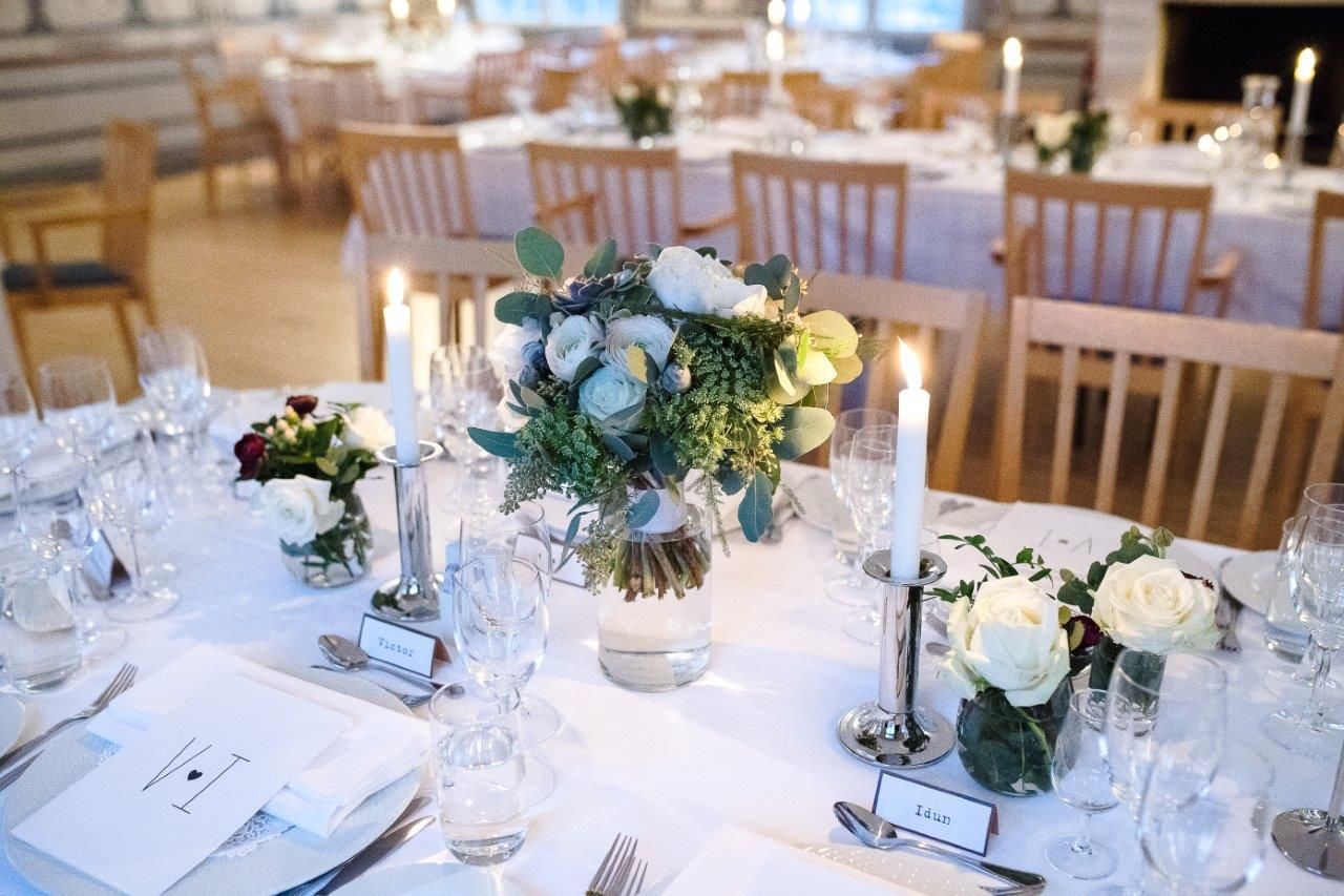 Bryllupsfestbord dekket med hvite duker, blomster, lys og kuverter.