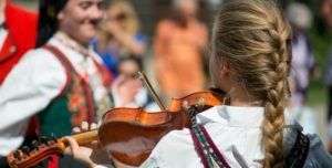 En person i tradisjonelle klær spiller fiolin utendørs, mens en annen person i folkeklær står i nærheten.