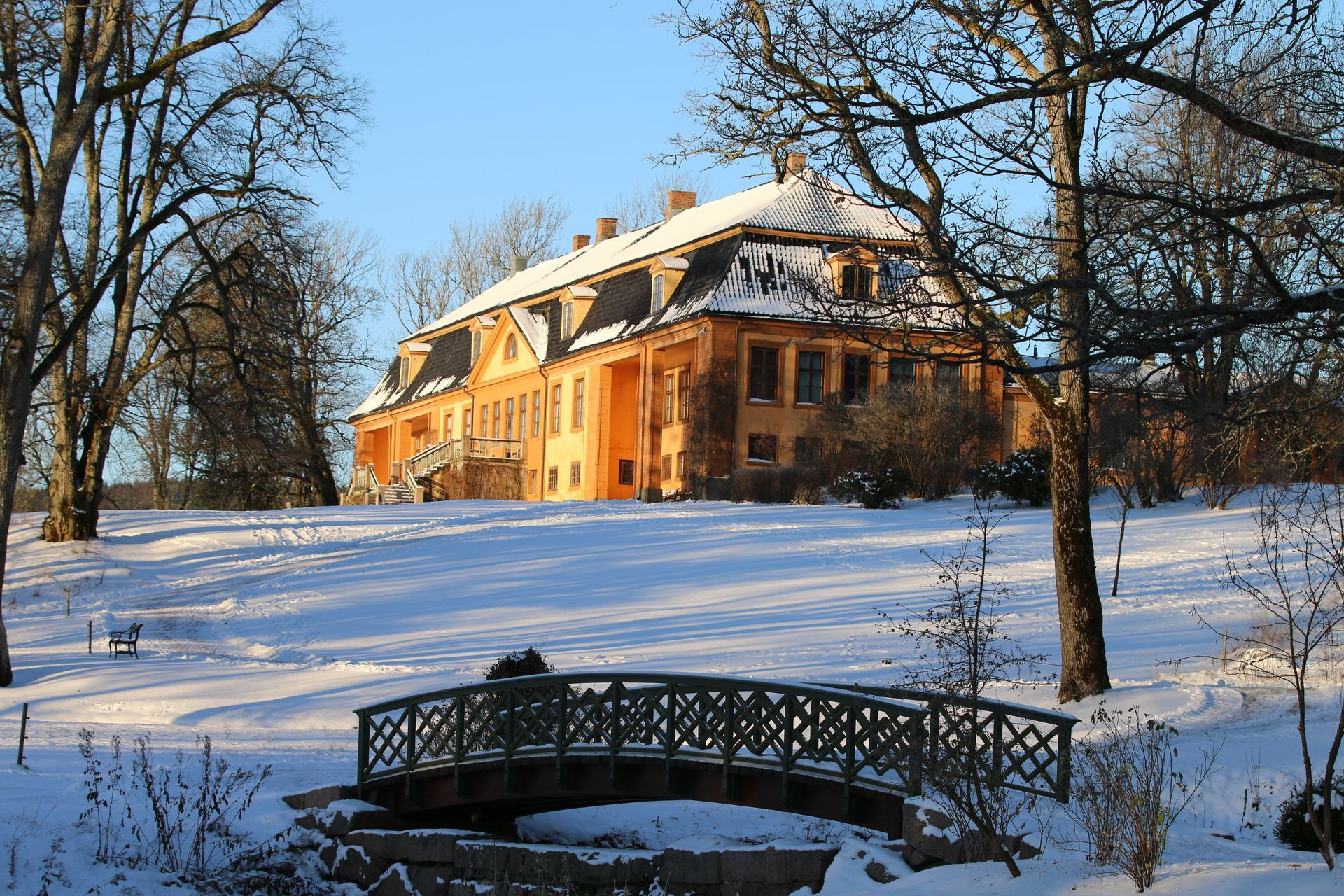Et oransje herregård med snødekt tak ligger bak en liten gangbro i et snødekt, solfylt landskap.
