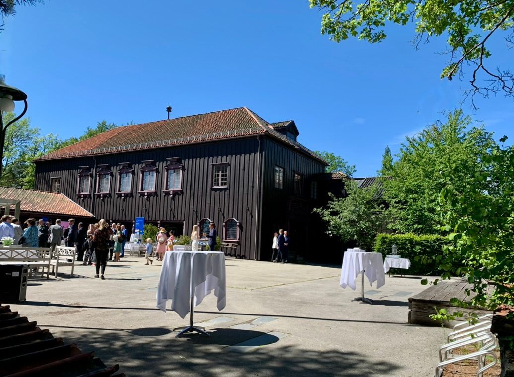 Utendørs gårdsplass med en mørk trebygning, bord med hvite duker og folk som omgås hverandre på en solrik dag.