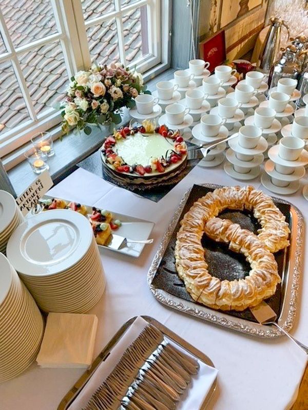 Buffetbord med kake, kringle, frukt, tekopper og bestikk plassert nær et vindu.