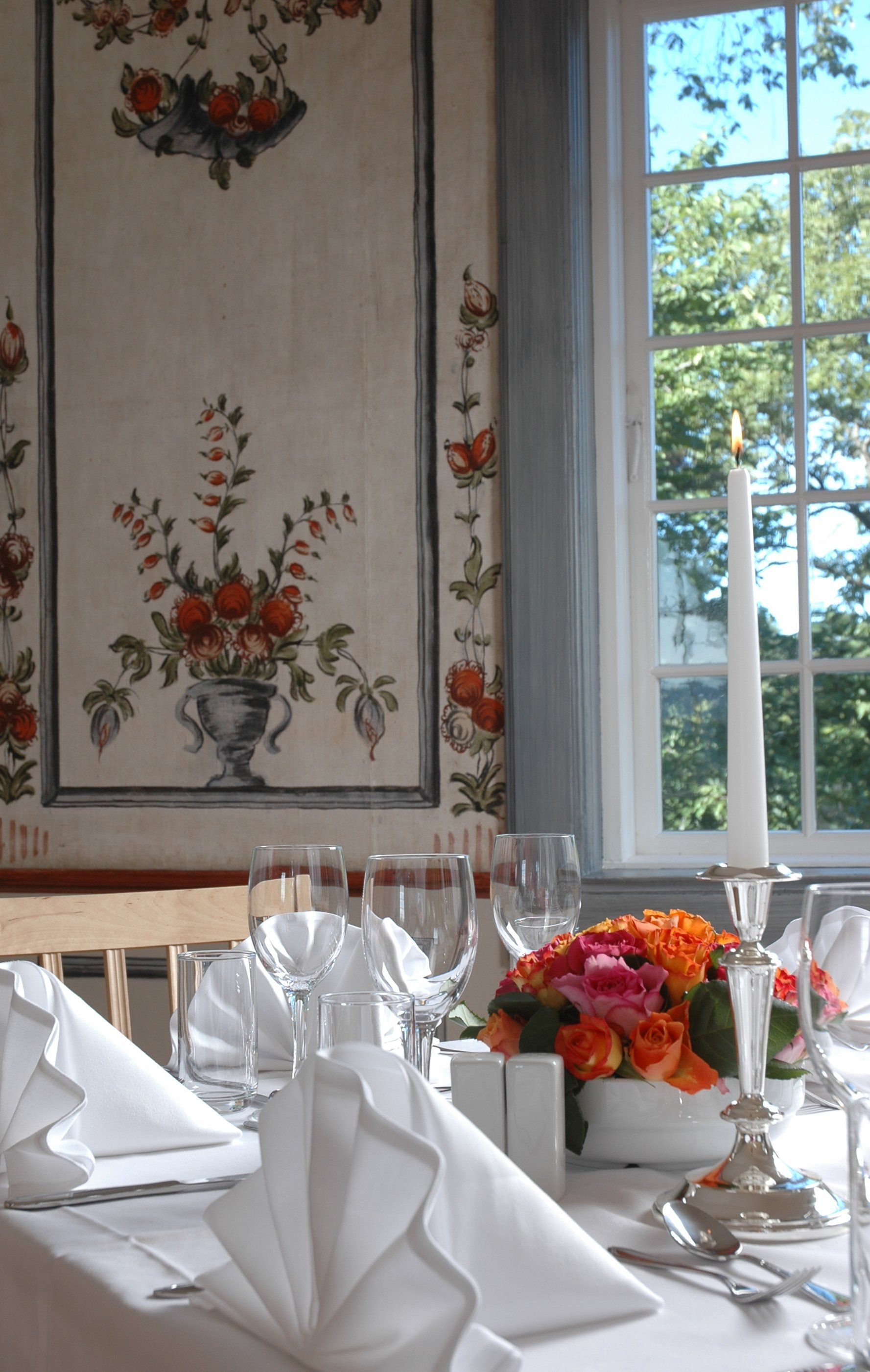 Formelt spisebord plassert nær et vindu med blomstertapet. Lyse blomster og et tent stearinlys er sentrert.