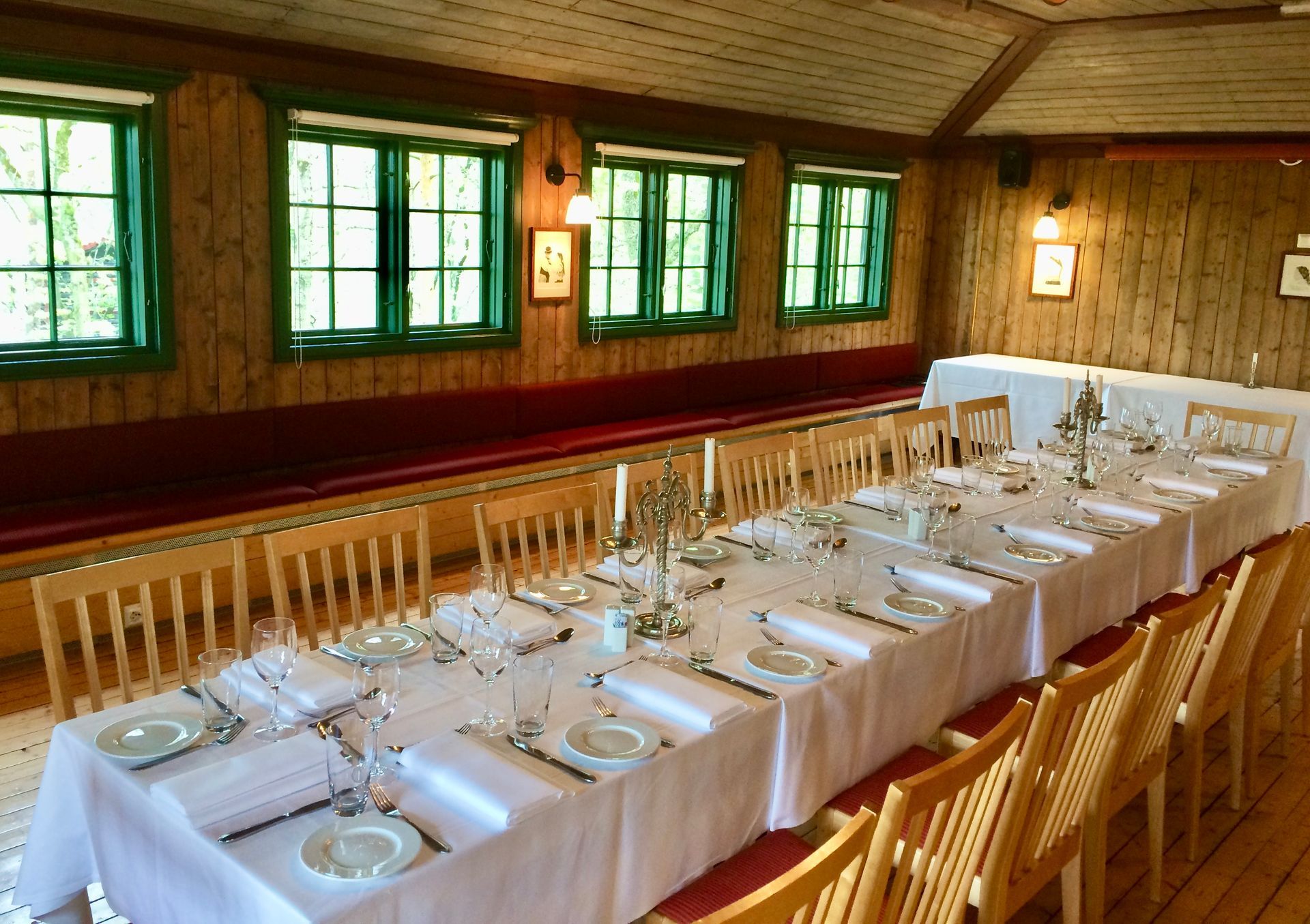 Langt spisebord dekket for et måltid i et trerom med grønne vinduer og en rød benk.