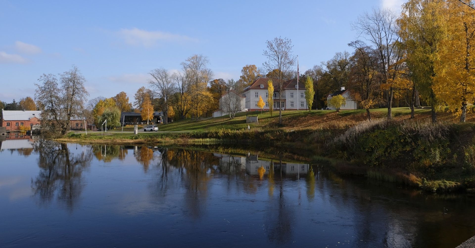 Et vannlegeme reflekterer bygninger og trær med høstgule blader under en blå himmel.