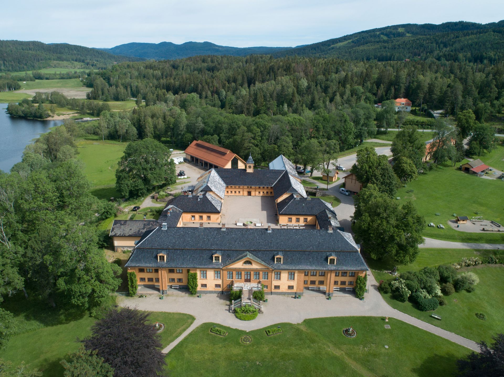 Luftfoto av et gult herregård med gårdsplass, omgitt av grønne åkre, skog og en innsjø.