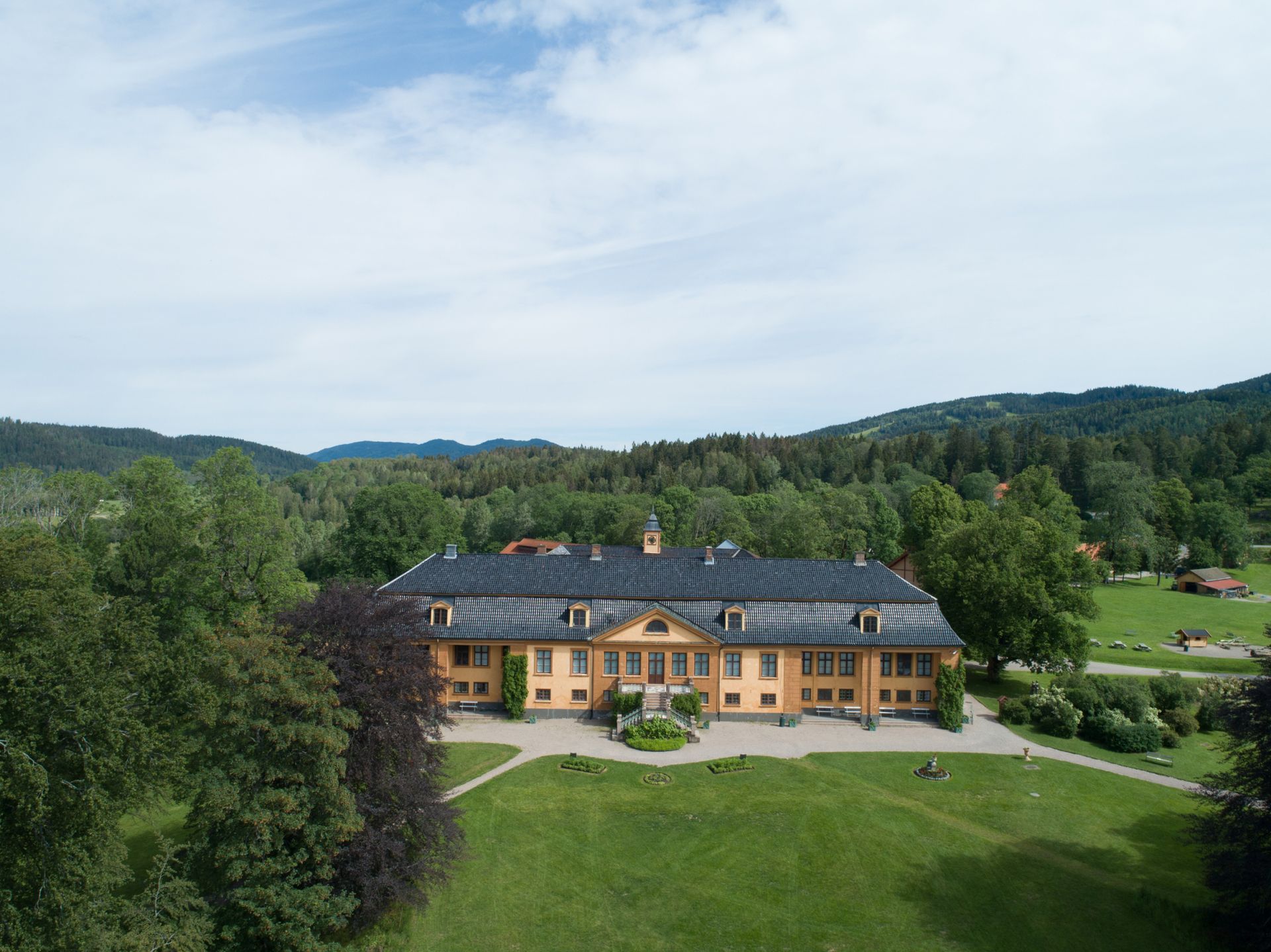 Gult herregård med mørkt tak på en gresskledd ås, omgitt av trær og et bakteppe av grønne åser.