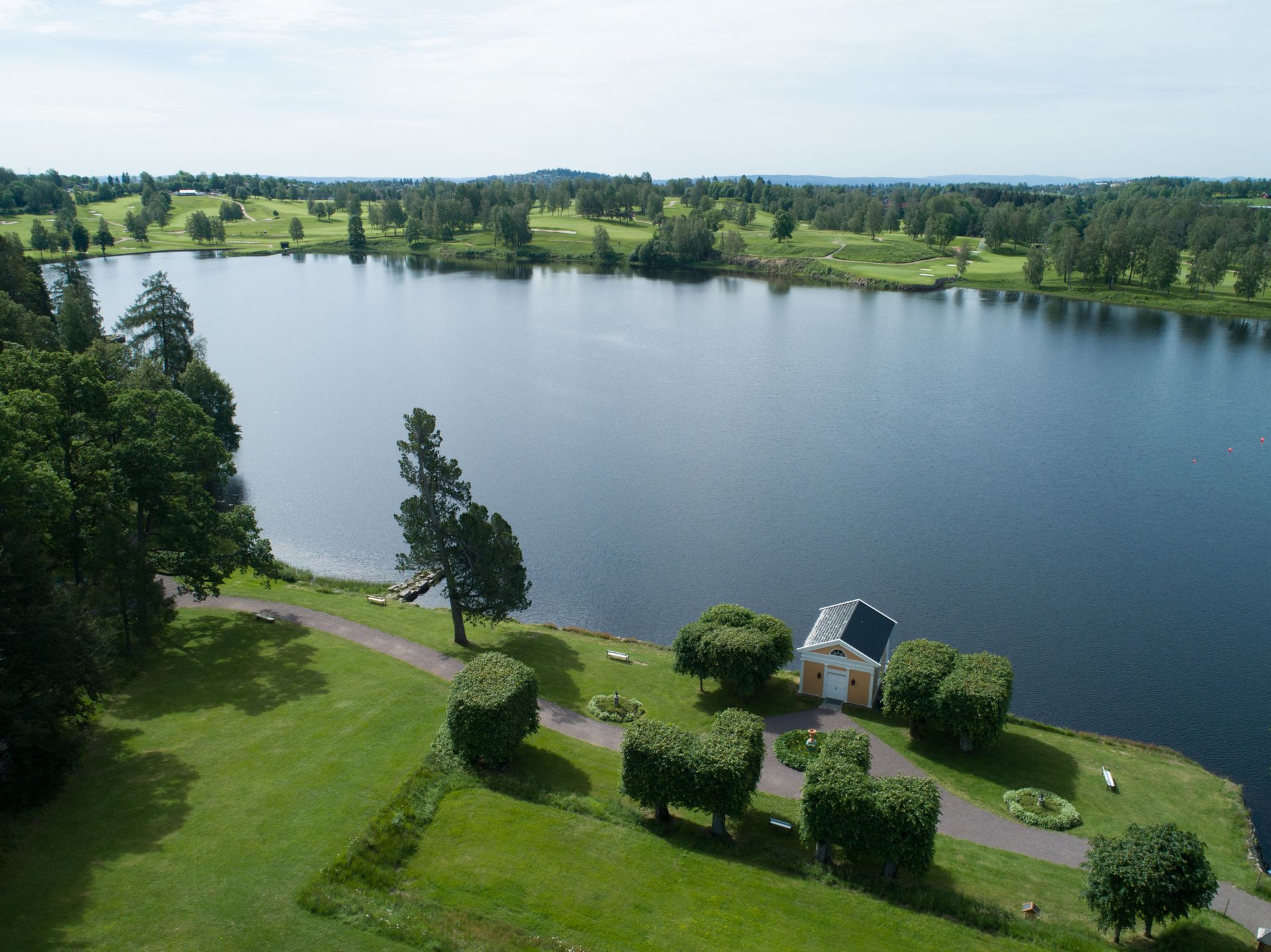 Utsikt over innsjøen med grønne plener, trær, en liten gul bygning og en golfbane i det fjerne.