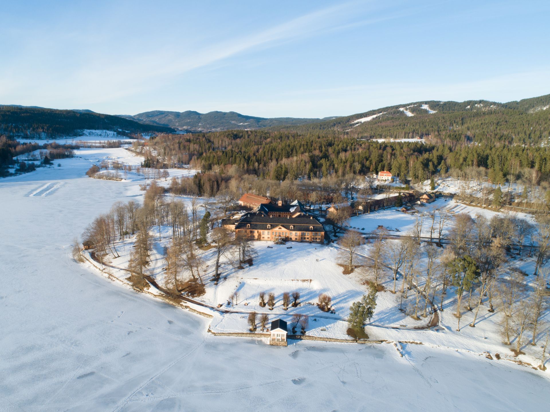 Luftfoto av et snødekt landskap med en stor bygning, omgitt av en frossen innsjø og skog.