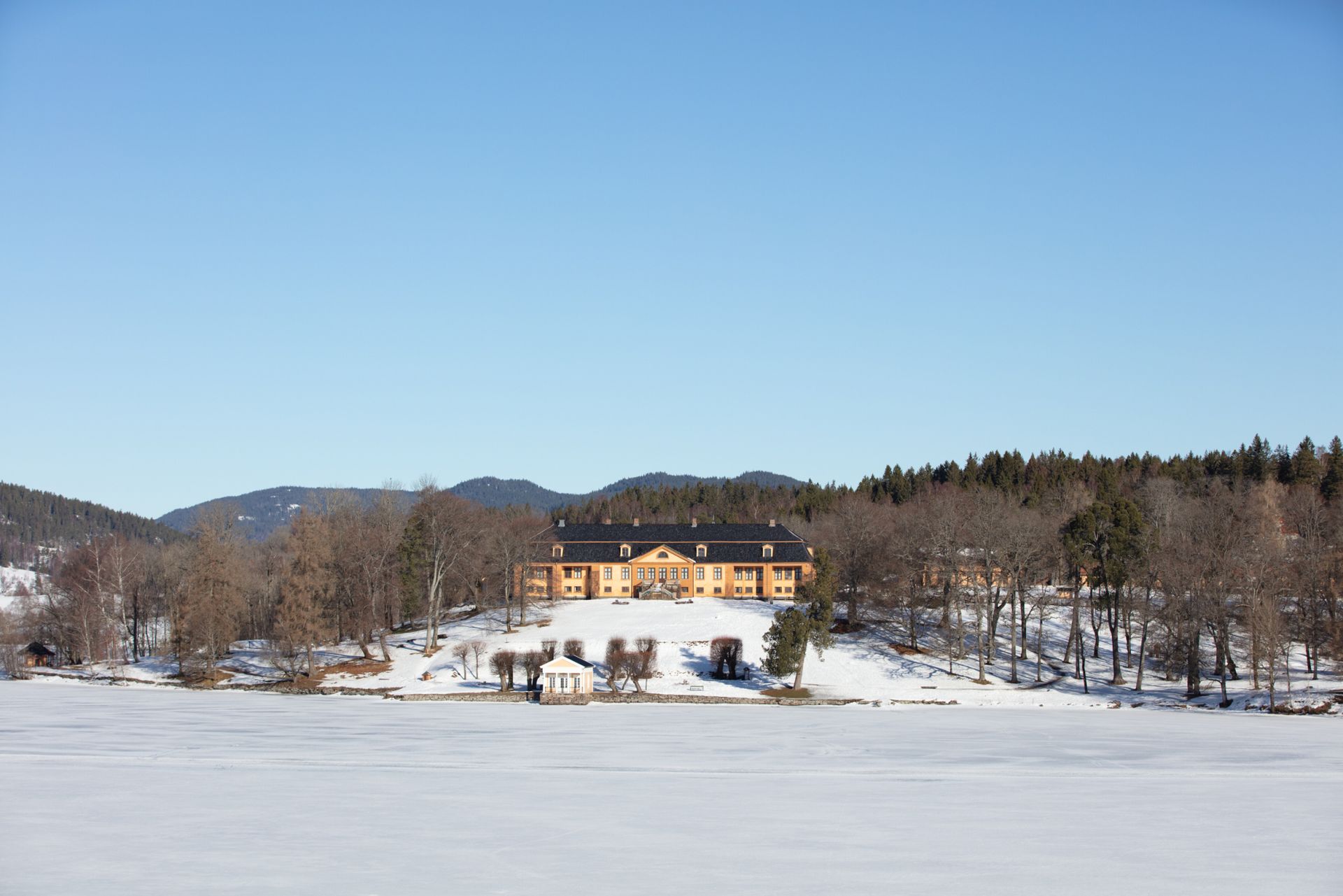 Gul bygning på snødekt ås ved en frossen innsjø, trær og blå himmel i bakgrunnen.
