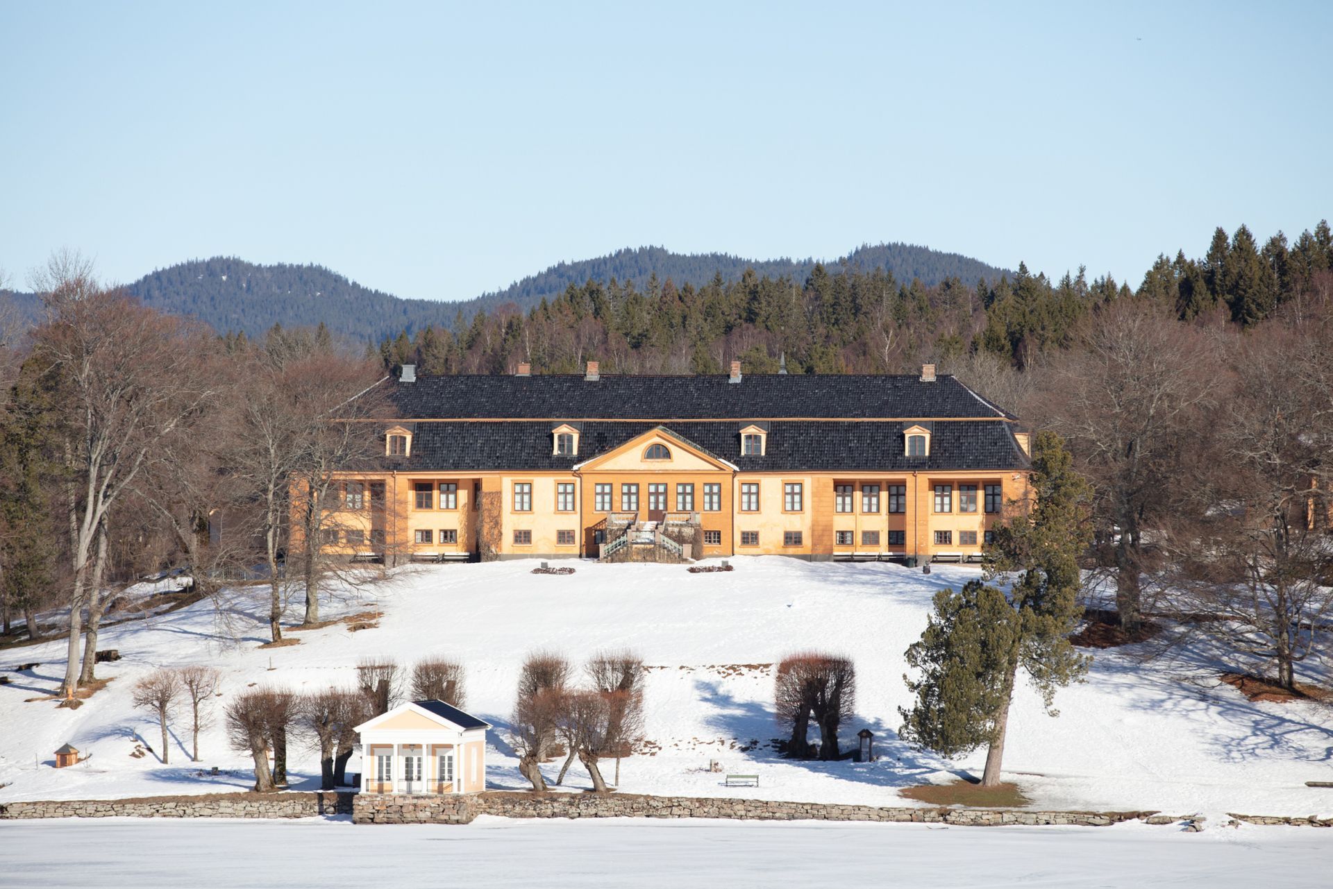 Gult herregård på en snødekt ås, omgitt av trær og en frossen innsjø, under en blå himmel.