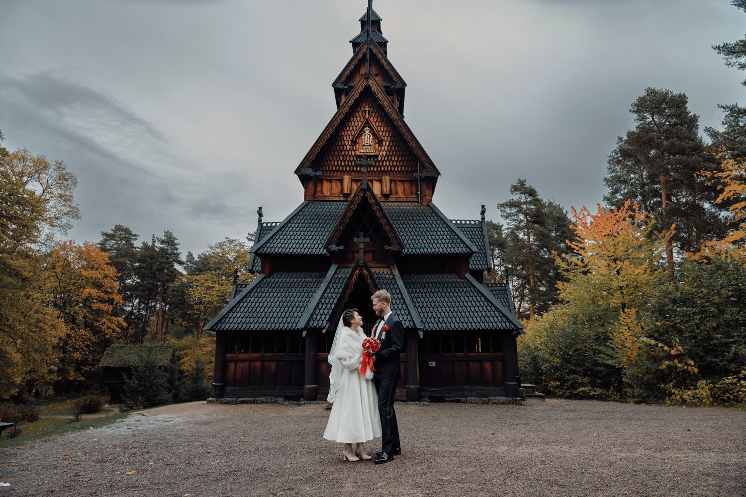 Par i brudekjole står foran en trekirke med et mørkt, spisst tak.