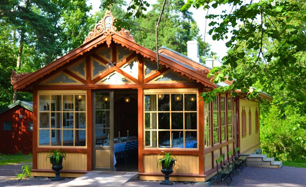 Trebygning med store vinduer og intrikate takdetaljer, omgitt av trær og grøntområder.