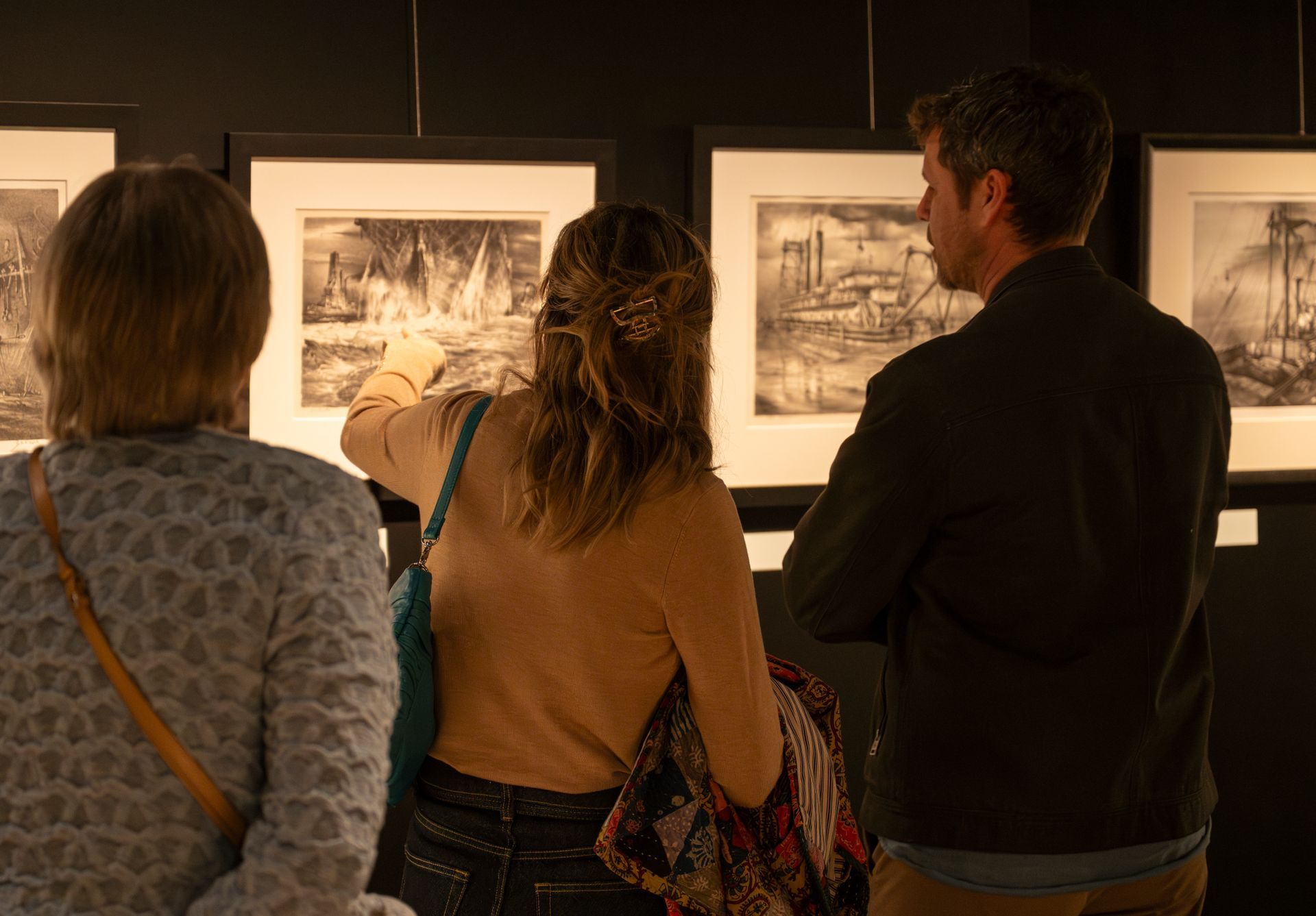 Folk ser på innrammet kunst på en gallerivegg; én person peker på et trykk.