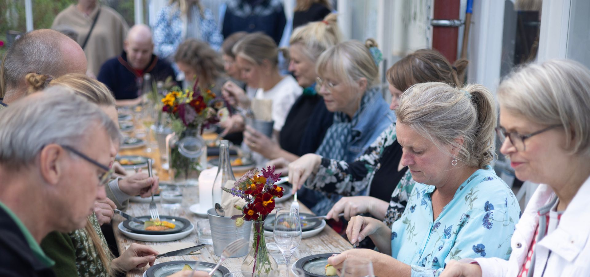 Et langt middagsbord dekket med tallerkener og blomster, hvor en gruppe mennesker sitter og spiser i et innendørs, naturlig opplyst område.