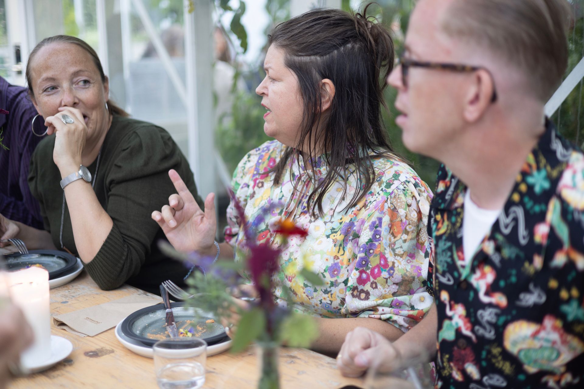 Folk omgås utendørs på en solrik sammenkomst, mens de holder drinker i nærheten av steintrapper og blomstrende busker.