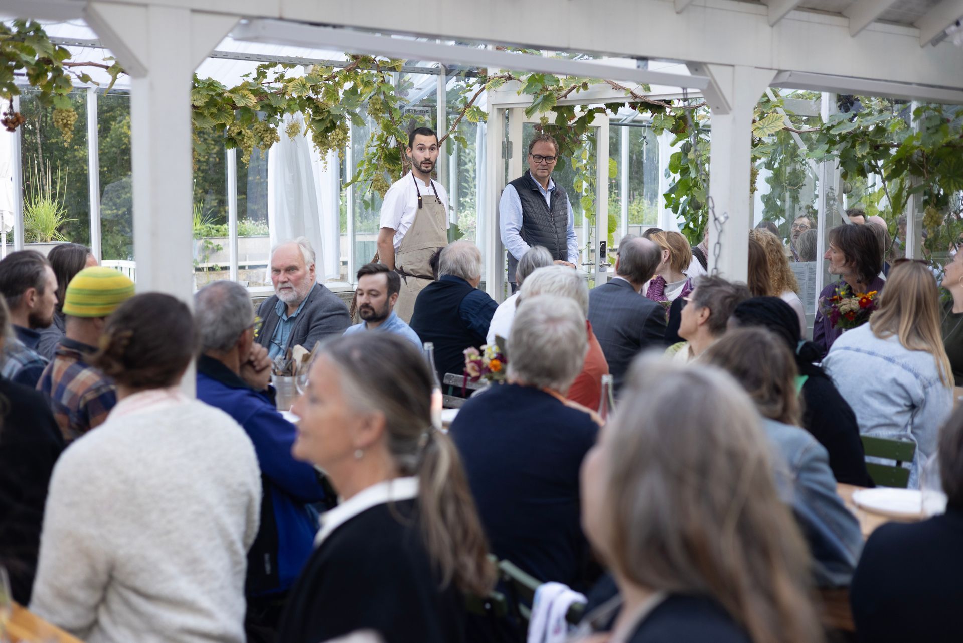Folk samles utendørs til et sosialt arrangement, med to personer i forgrunnen som snakker og holder vinglass.