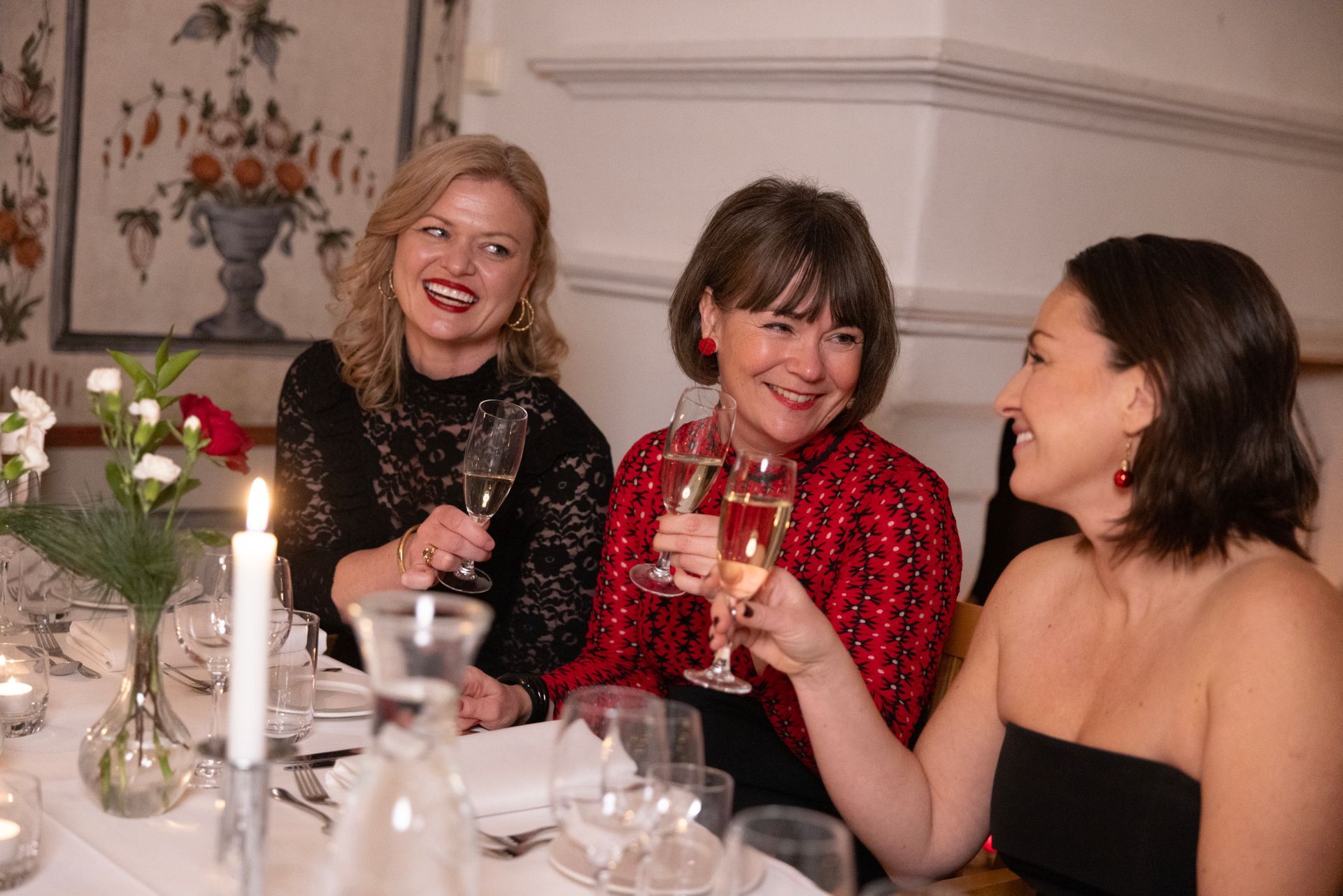 Tre kvinner klinker champagneglass under en festmiddag. En i svart blonde, en annen i rødt og en i en svart stroppeløs topp, alle smilende.
