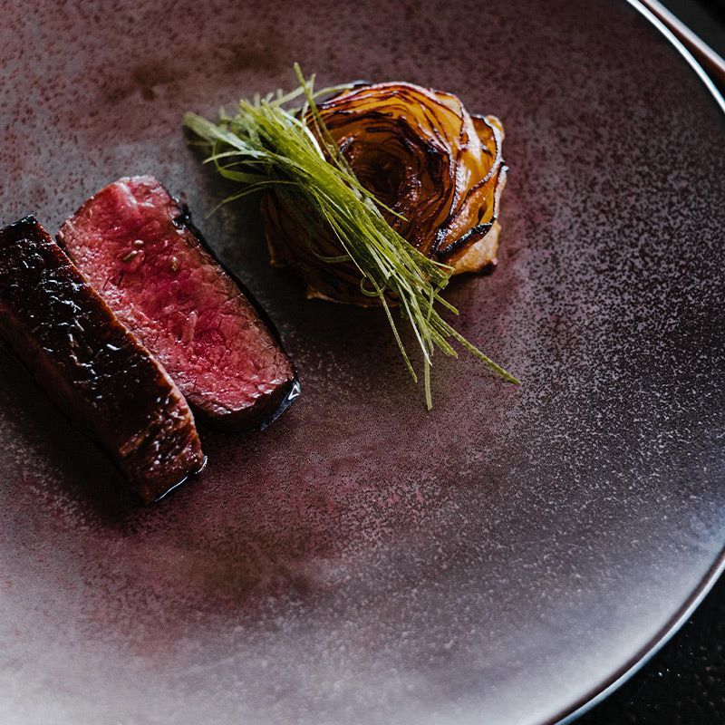 En sjeldent stekt biff servert med en forkullet løkblomst og frisk gressløk på en mørk, teksturert tallerken.