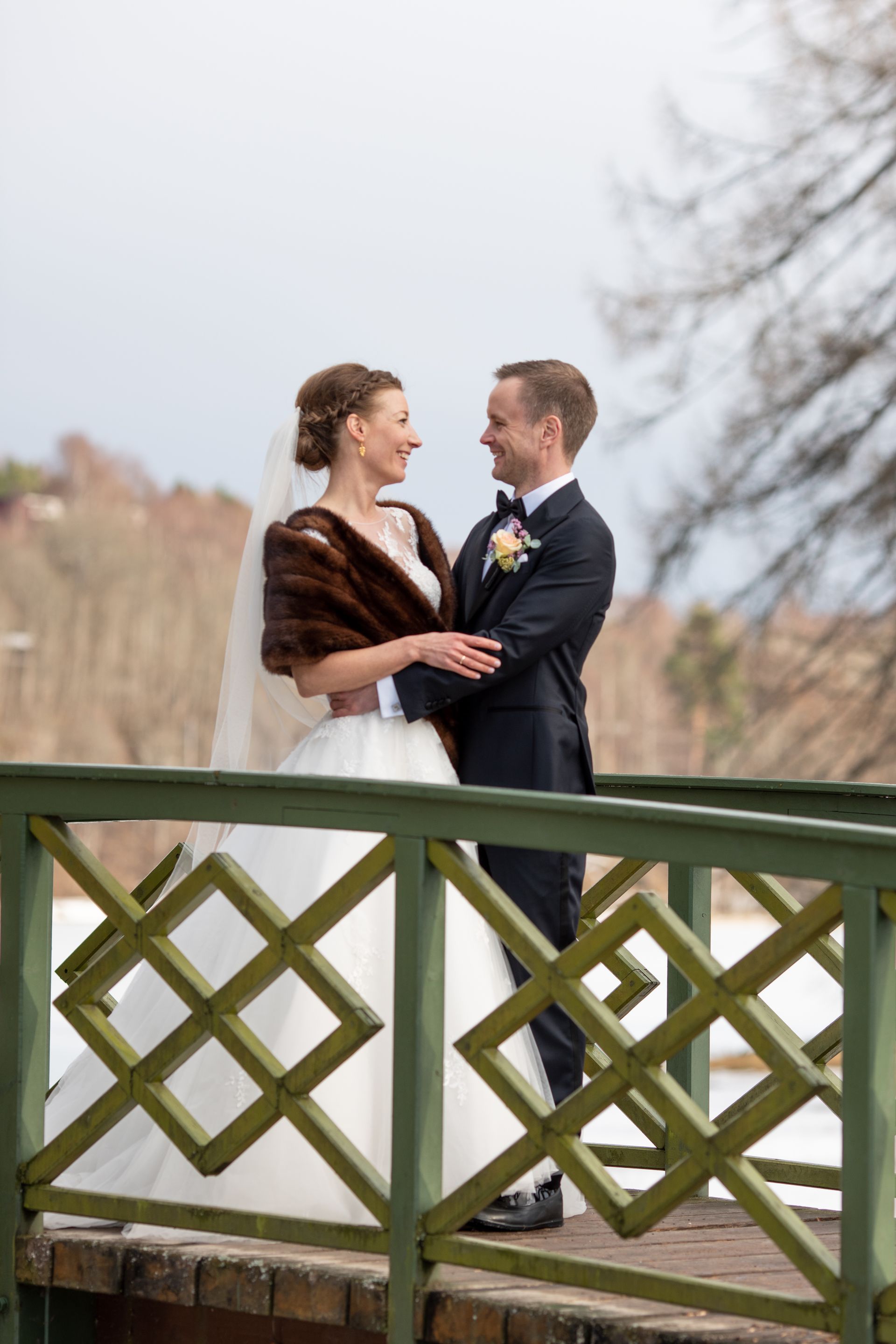 Brud og brudgom smiler til hverandre, stående på en grønn bro; bruden har på seg et pelssjal.