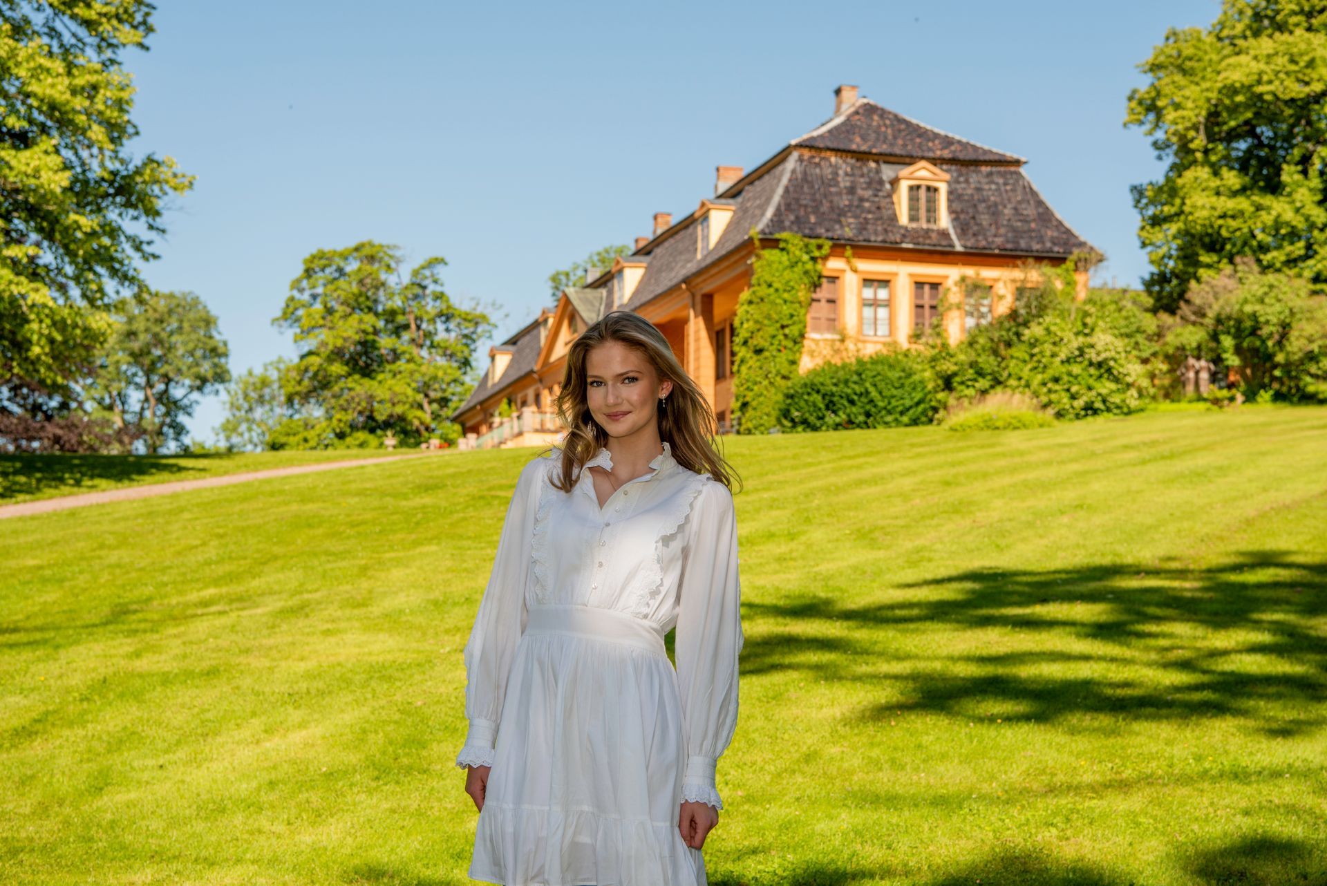 Kvinne i hvit kjole står på en gresskledd ås foran et gult hus på en solskinnsdag.