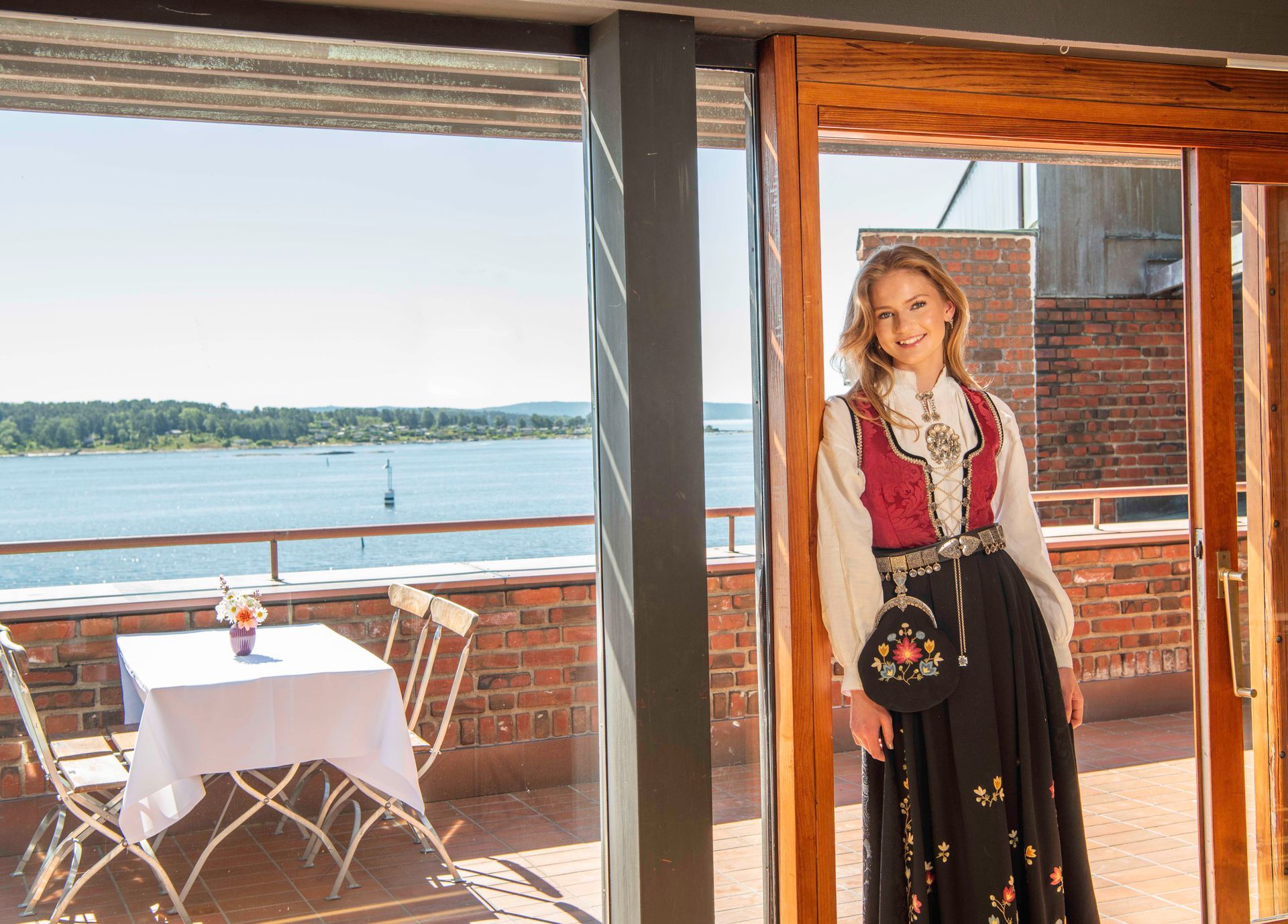 Konfirmant i bunad står ved åpne dører med utsikt over et vann.