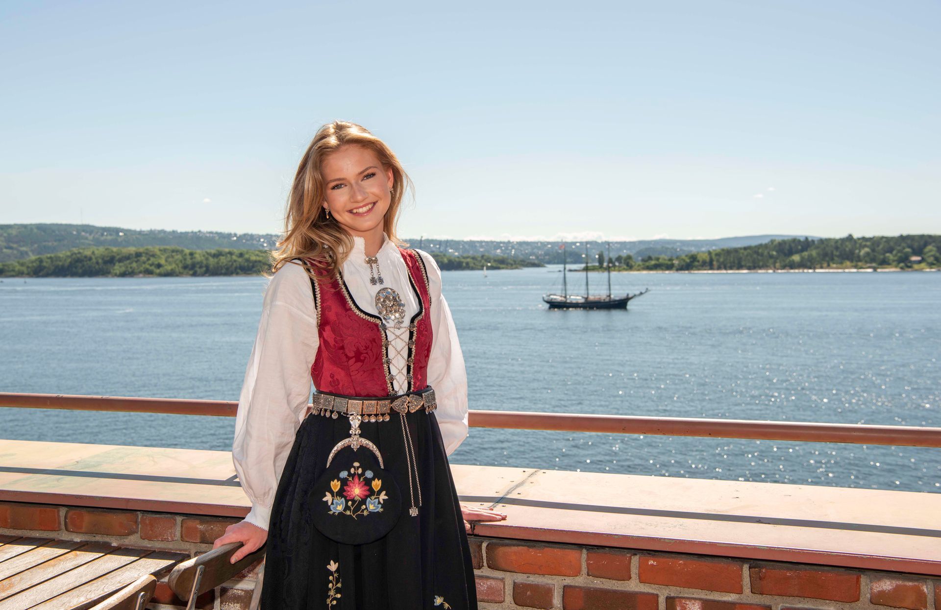 Konfirmant i bunad poserer på en balkong med utsikt over vannet, med en seilbåt i det fjerne.