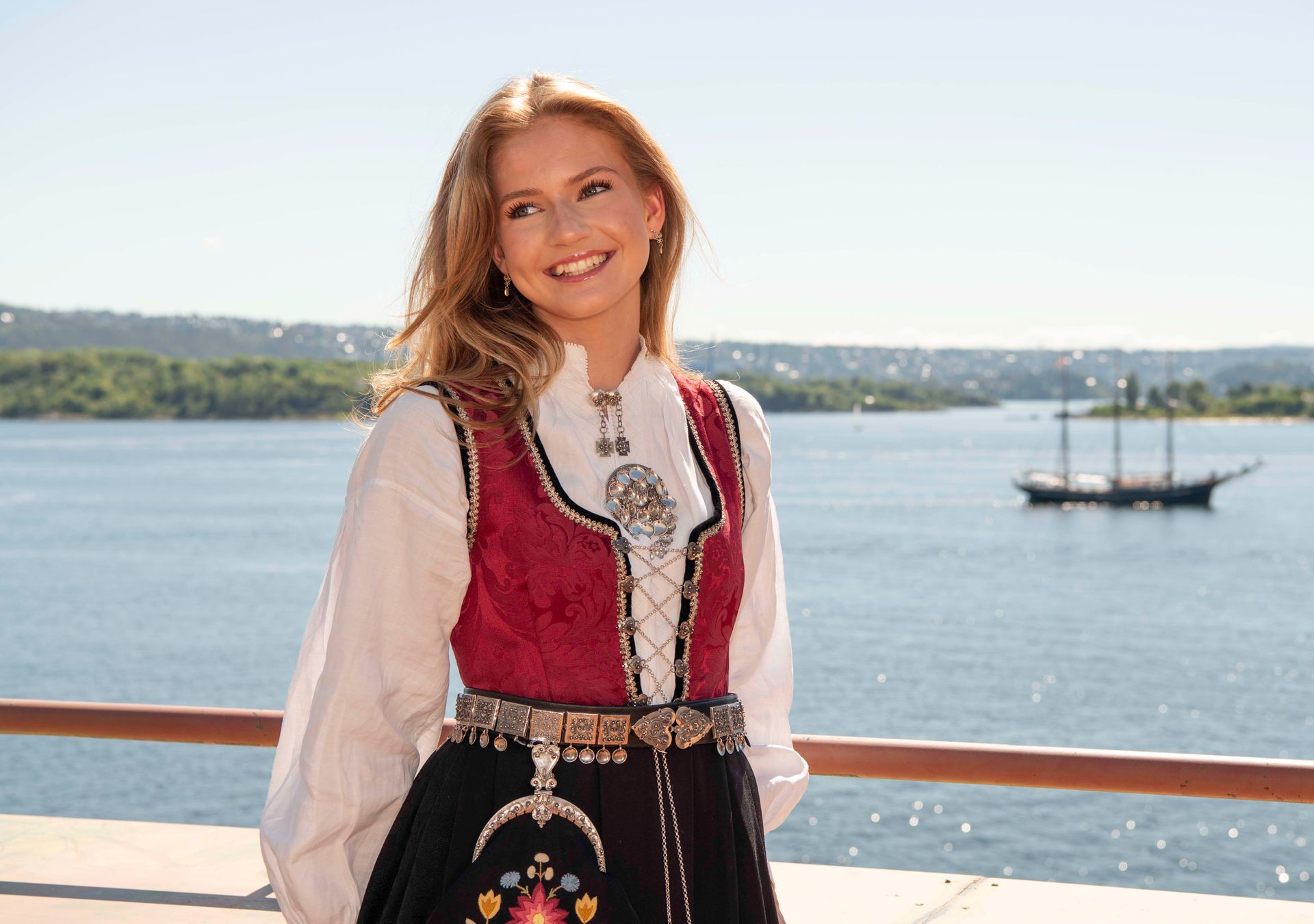 Kvinne i bunad smiler, poserer på en balkong med utsikt over havet.