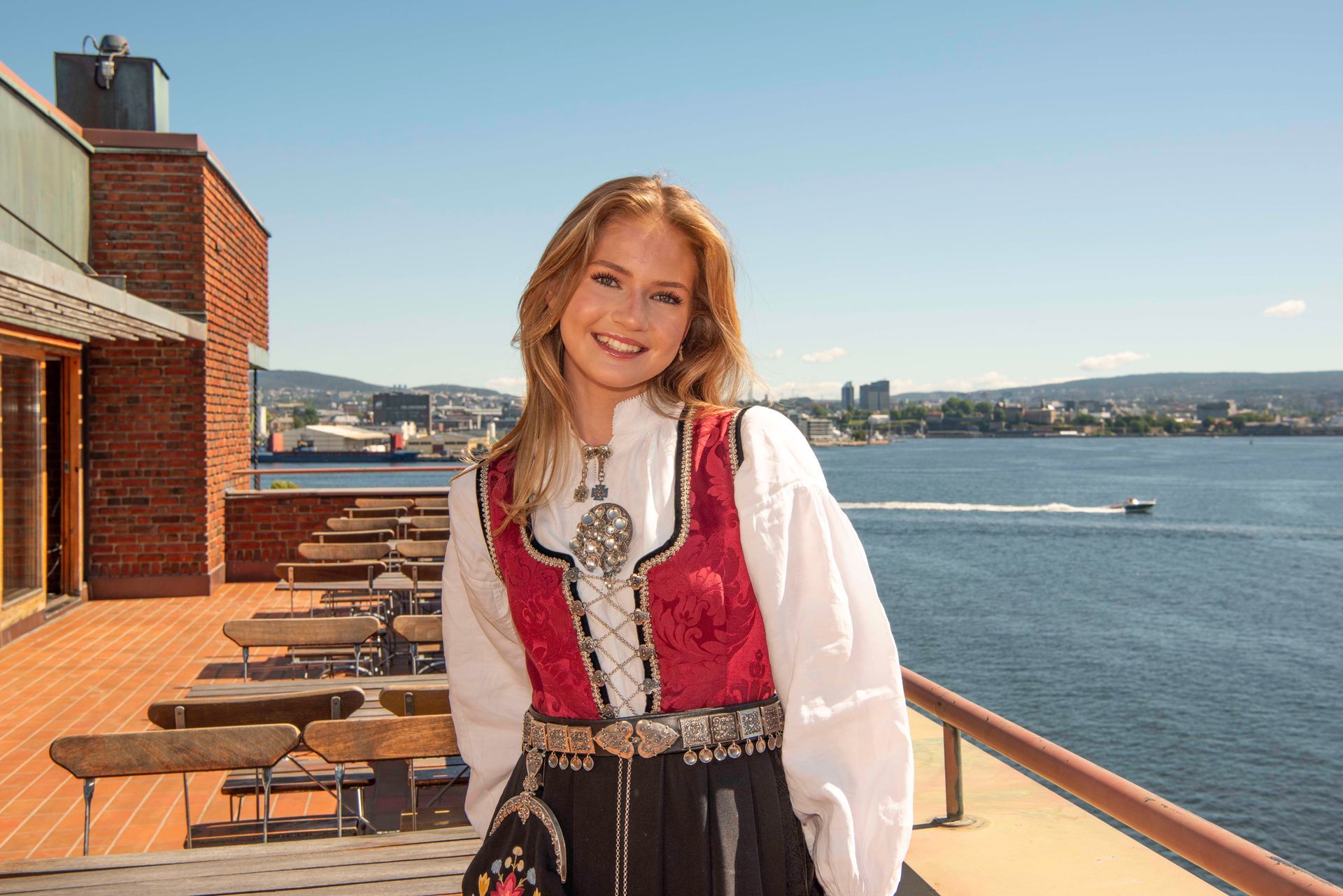 Konfirmant i bunad smiler på en balkong med utsikt over en bukt.