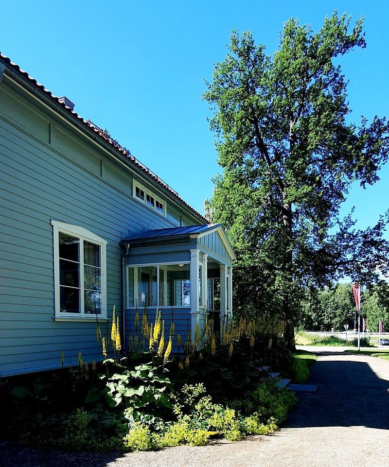 Lyseblått hus med veranda, frodig grønt løvverk og et tre mot en klar blå himmel.