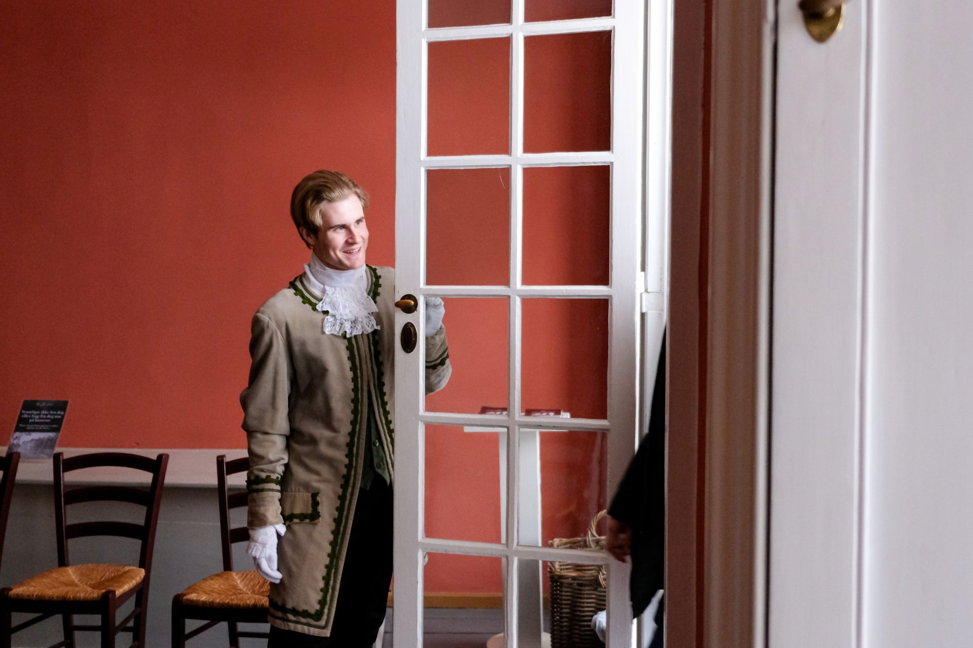 En mann i periodekostyme smiler og holder en dør med hvit innrammet dør åpen. En rød vegg og stoler i bakgrunnen.