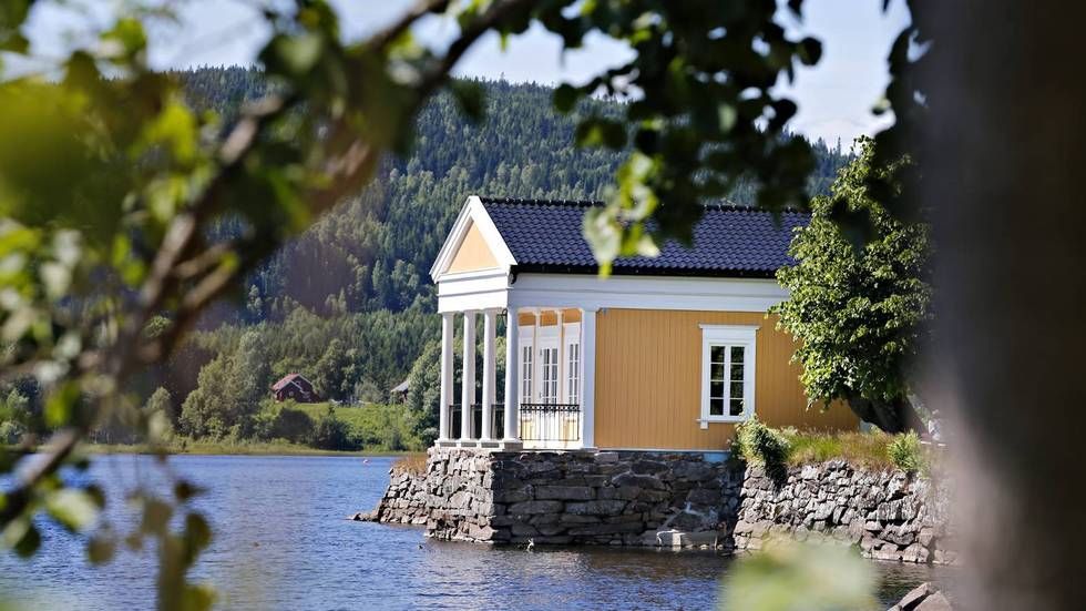 En gul paviljong i nyklassisistisk stil med hvite søyler står på en steinsokkel ved kanten av en rolig, blå innsjø.