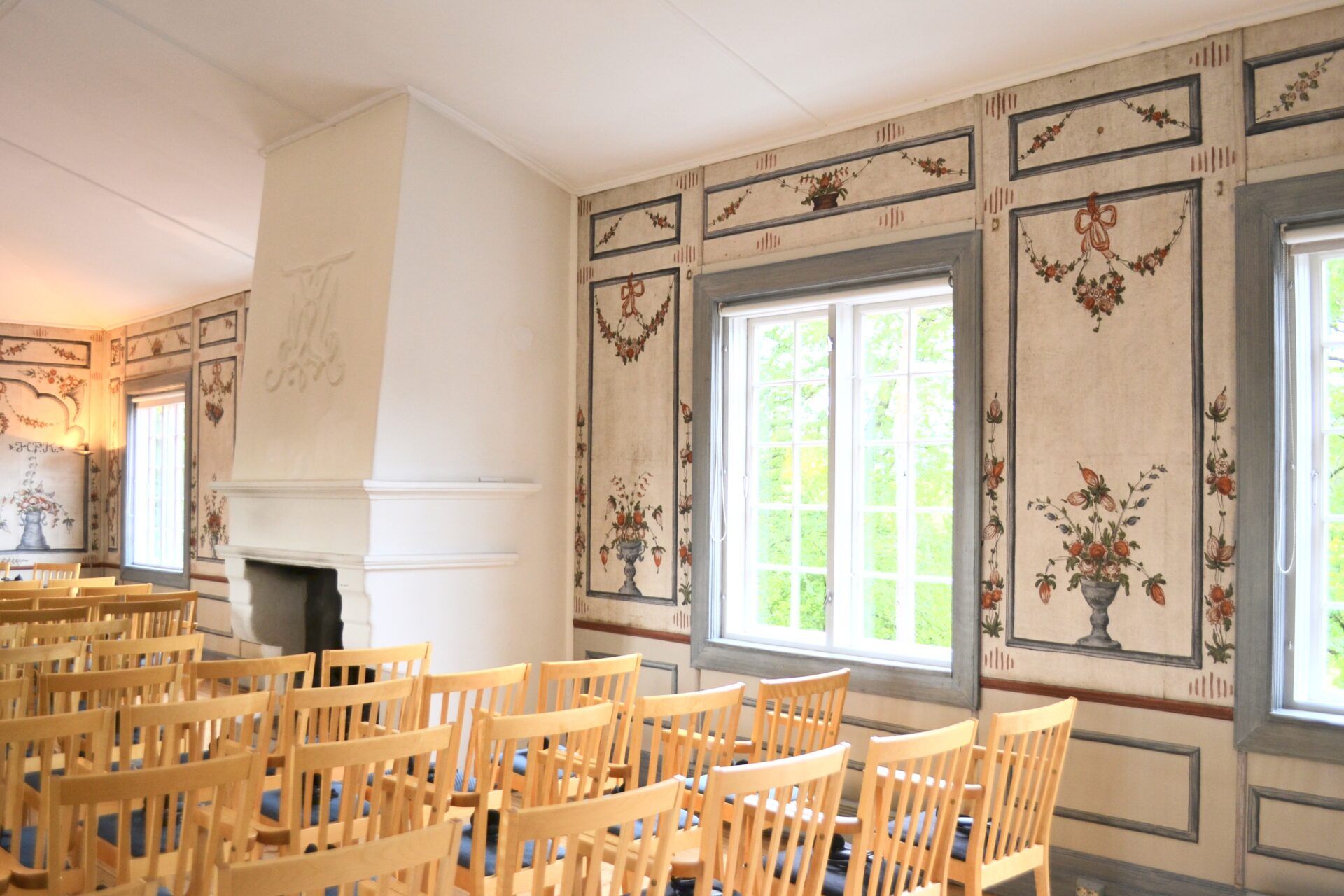 Et rom med rekker av trestoler vender mot vinduer og dekorerte vegger. En hvit peis er til venstre.