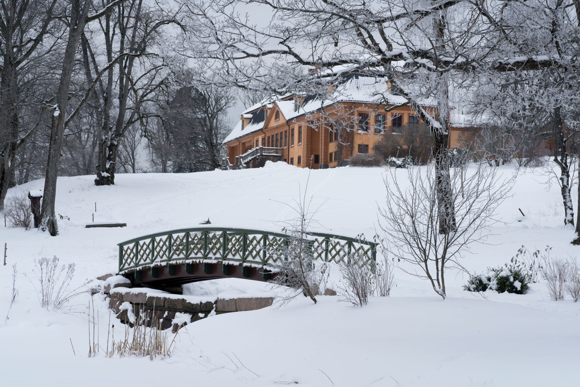 Snødekt landskap med en bro over en bekk, som fører til et gult hus på en ås.