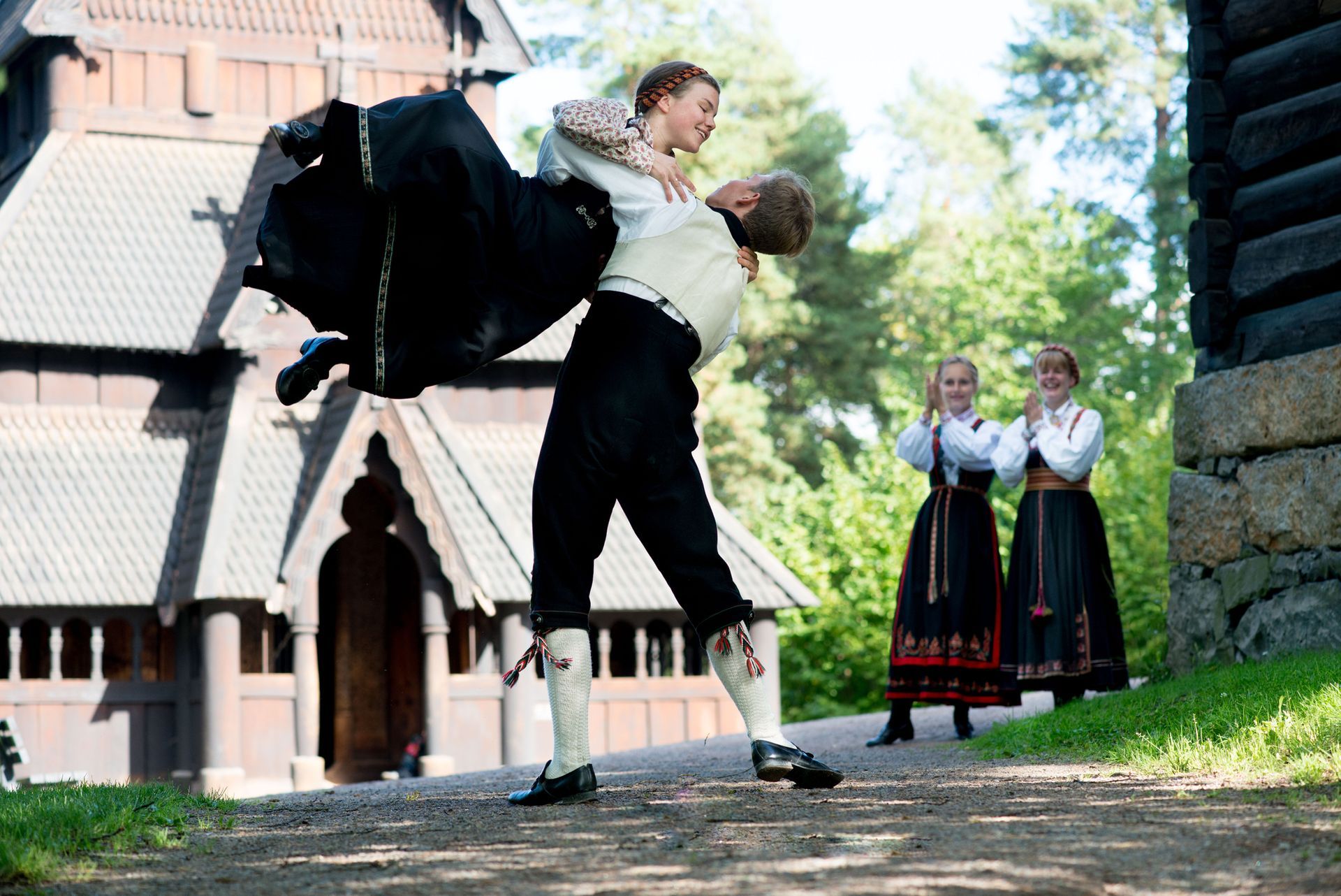 Mann løfter kvinne som danser utendørs, andre dansere ser på i nærheten av en trekirke.