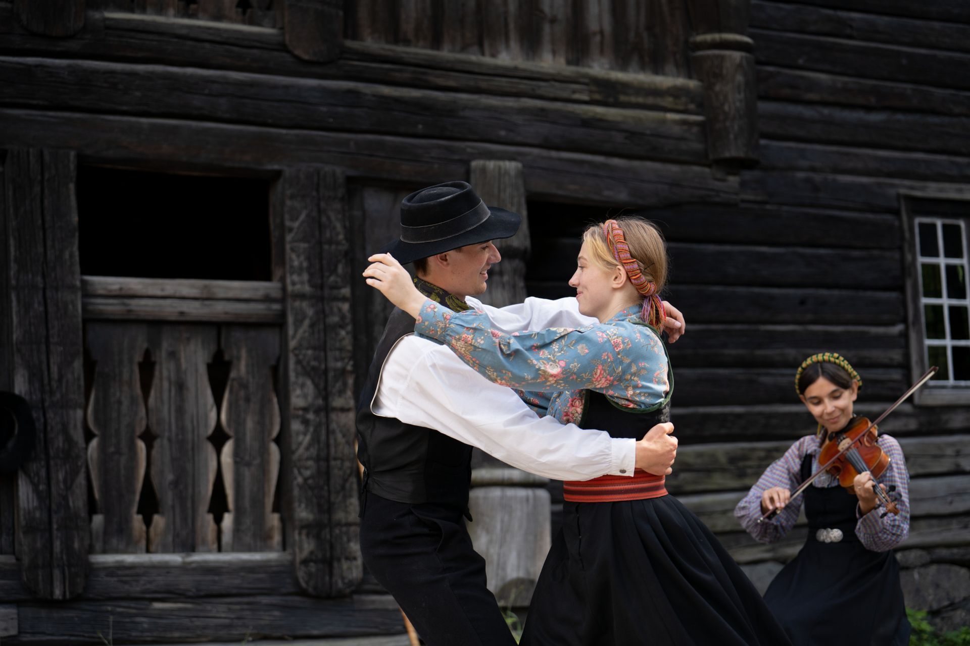 Pardanser i tradisjonelle klær; en musiker spiller fiolin foran en trebygning.
