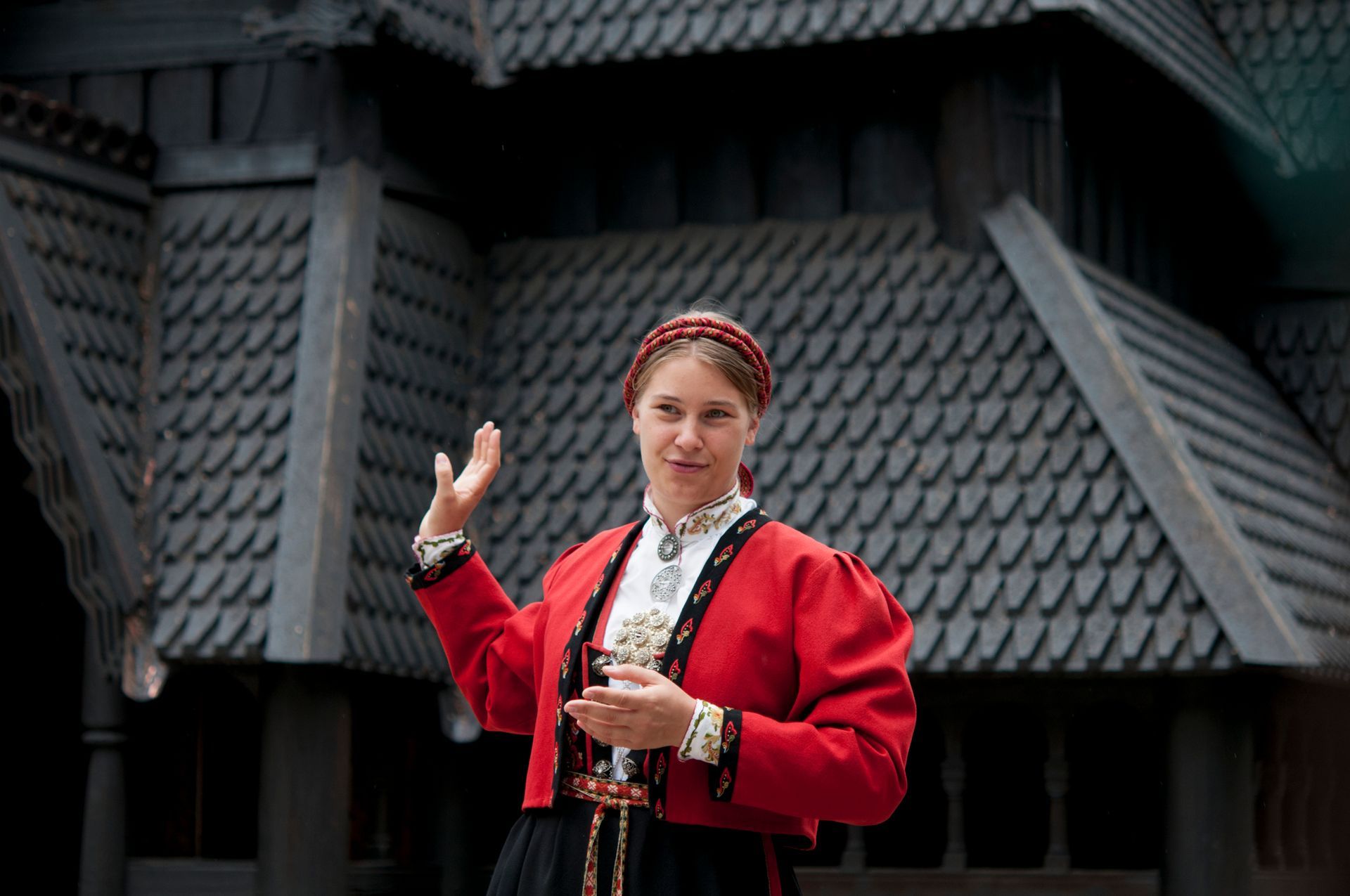 Kvinne i bunad gestikulerer foran en mørk stavkirke. 