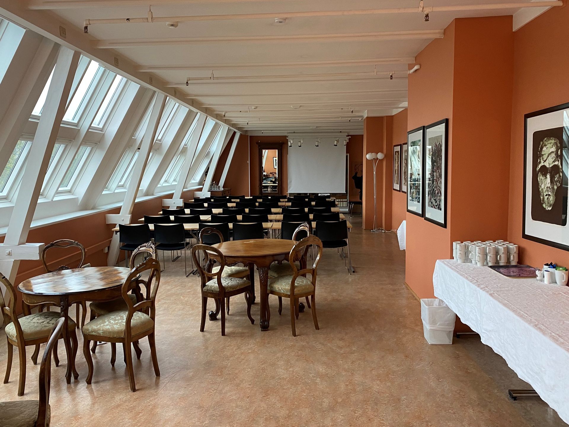 Konferanserom med vinklede vinduer, bord, stoler, projektorlerret og oransje vegger.
