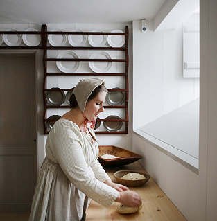 Kvinne i historiske klær elter deig på et kjøkken med trehyller med tallerkener.