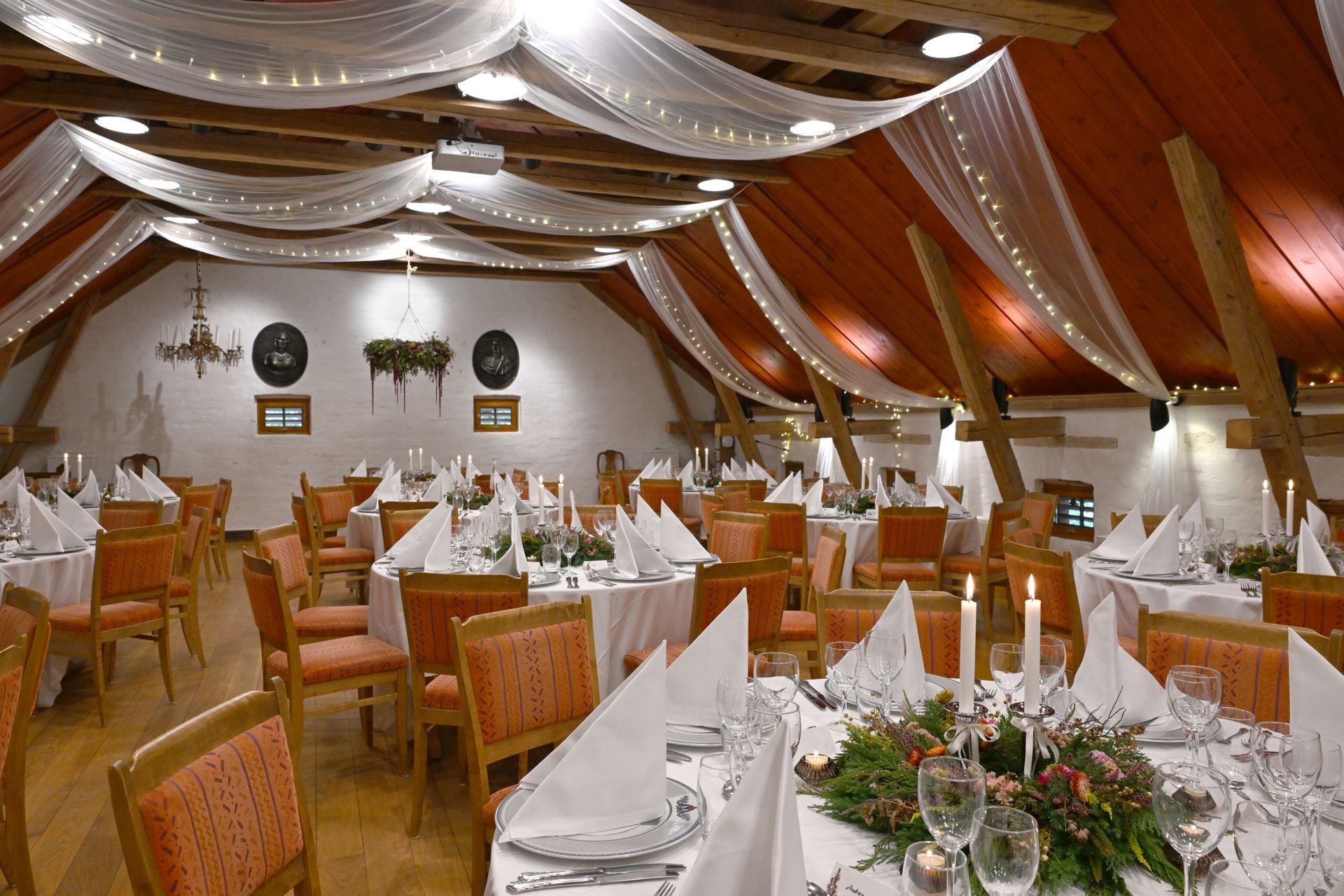 Formell spisesal med runde bord dekket til bankett, under drapert hvitt stoff og lyslenker.