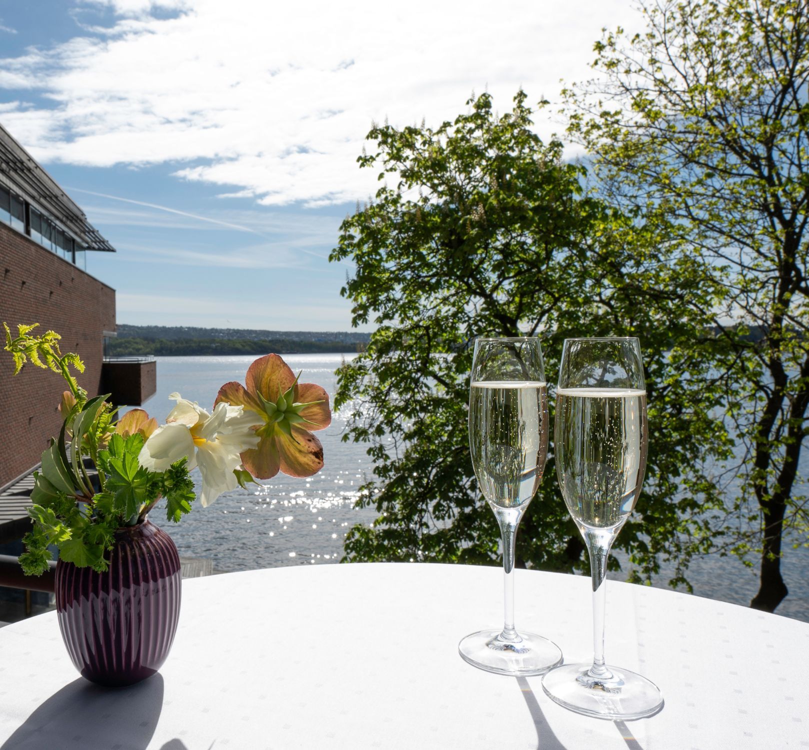 To champagnefløyter på en hvit duk med utsikt over innsjøen, blomster i en vase og en murbygning.
