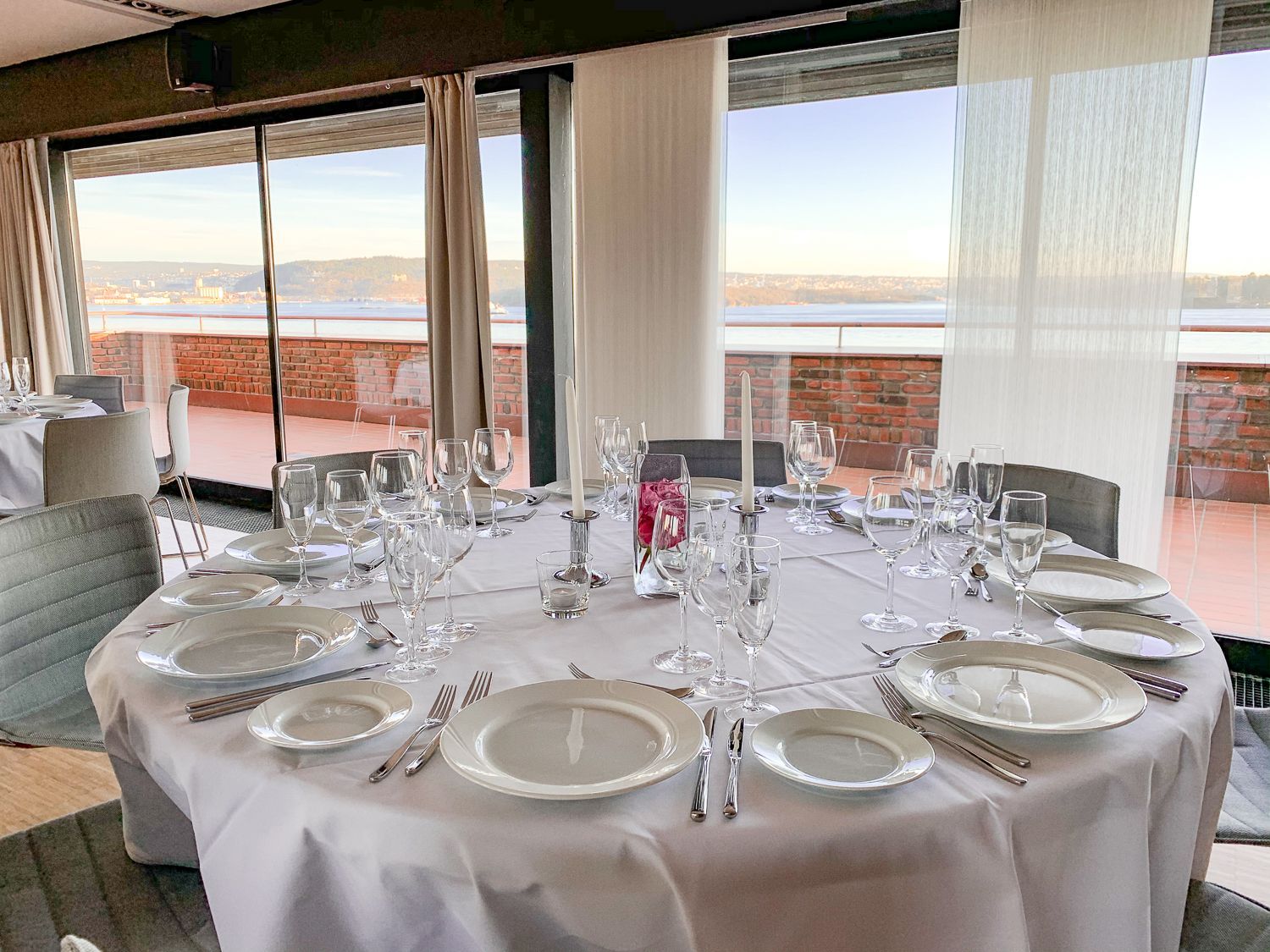 Rundt bord dekket for et måltid med tallerkener, glass og sølvtøy, med utsikt over et bybilde.