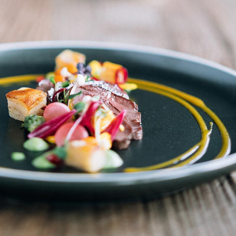 En gourmetrett med skiver av kjøtt, terninger av pynt og røde kronblader arrangert på en mørk tallerken med en virvel av gul saus.