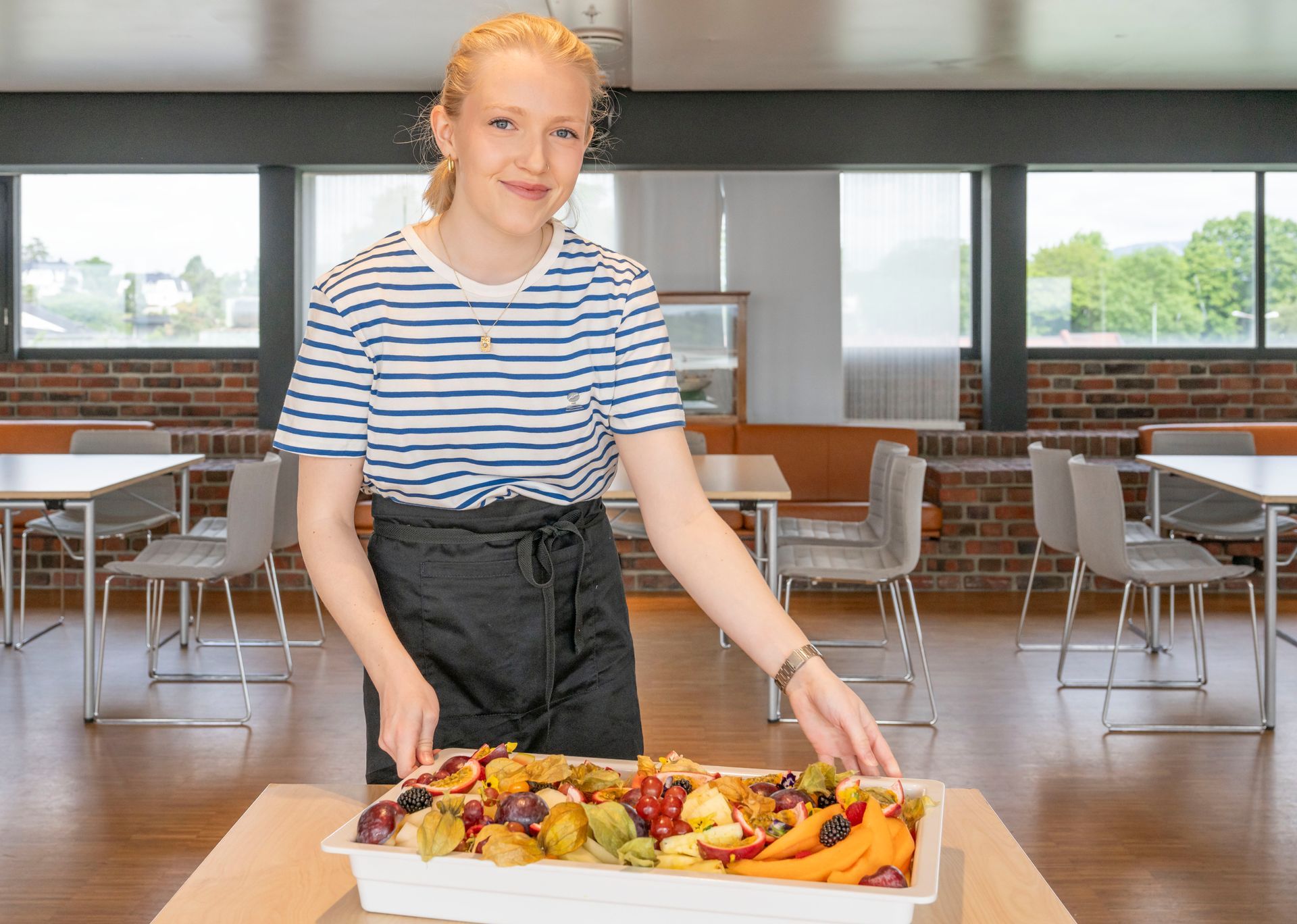 Kvinne i stripete skjorte som holder et brett med assortert frukt i en kantine.