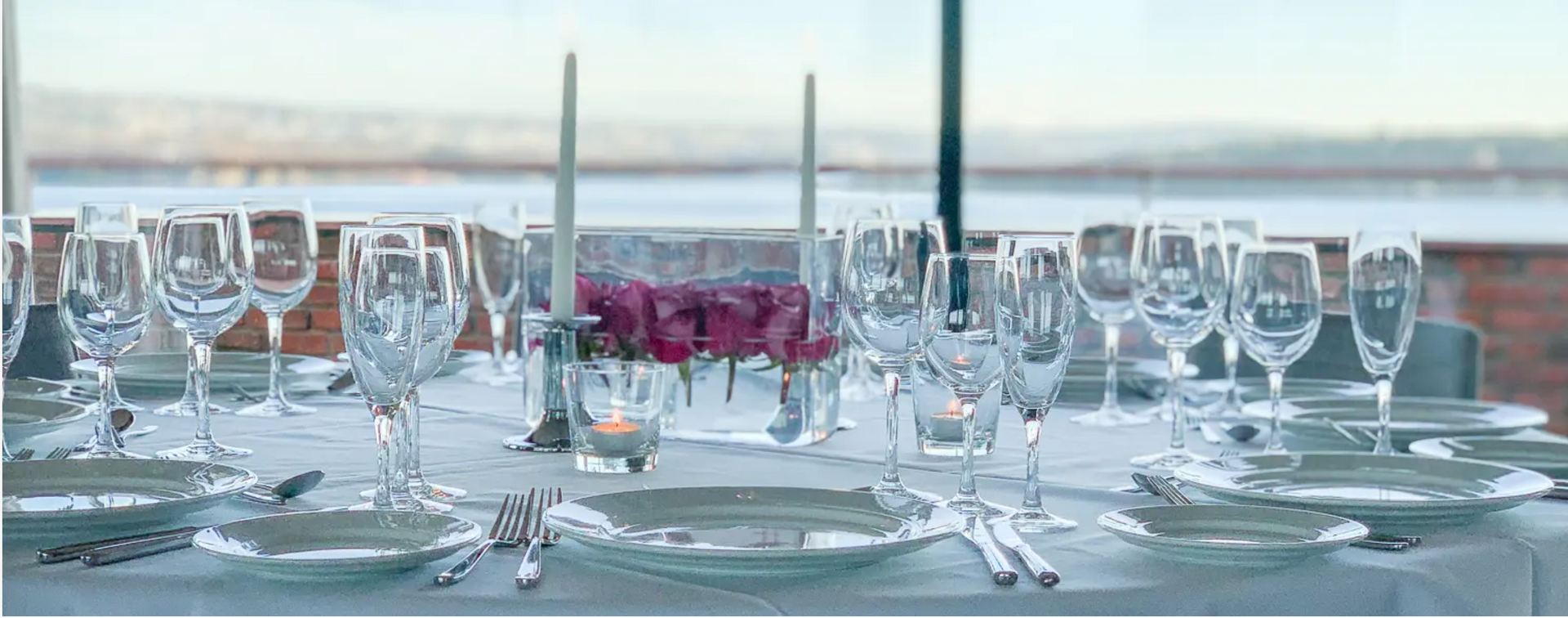 Formelt spisebord med glass, tallerkener, sølvtøy, stearinlys og blomsterdekorasjon, med utsikt over vannet.