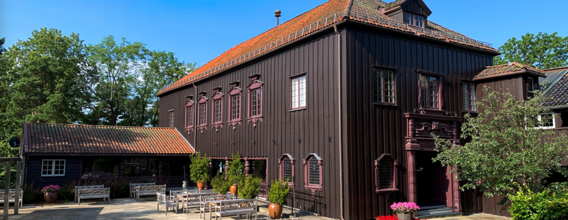 En mørk trebygning med rødt tegltak og rosa vindusrammer, bord og stoler står utenfor.