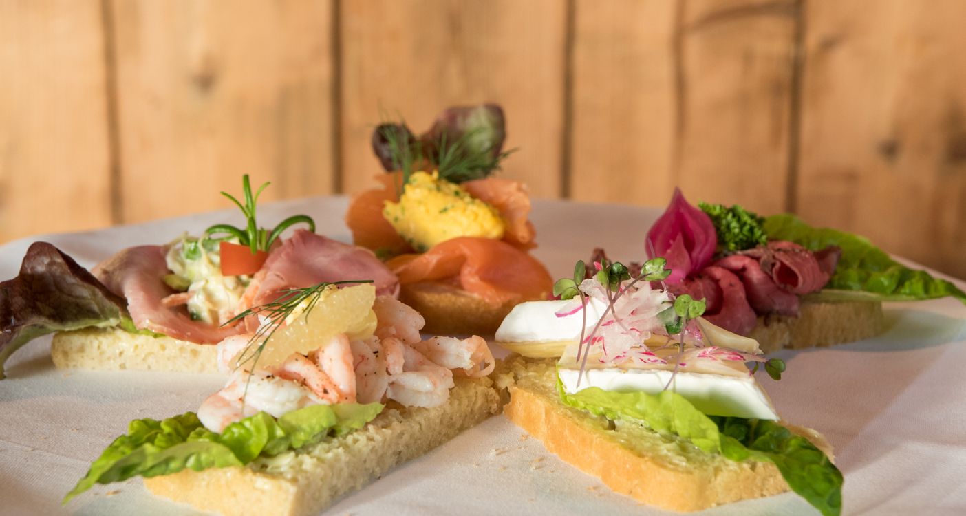 Et fat med fire assorterte forretter på ristede brødskiver, med toppinger som reker, laks og friske grønnsaker.