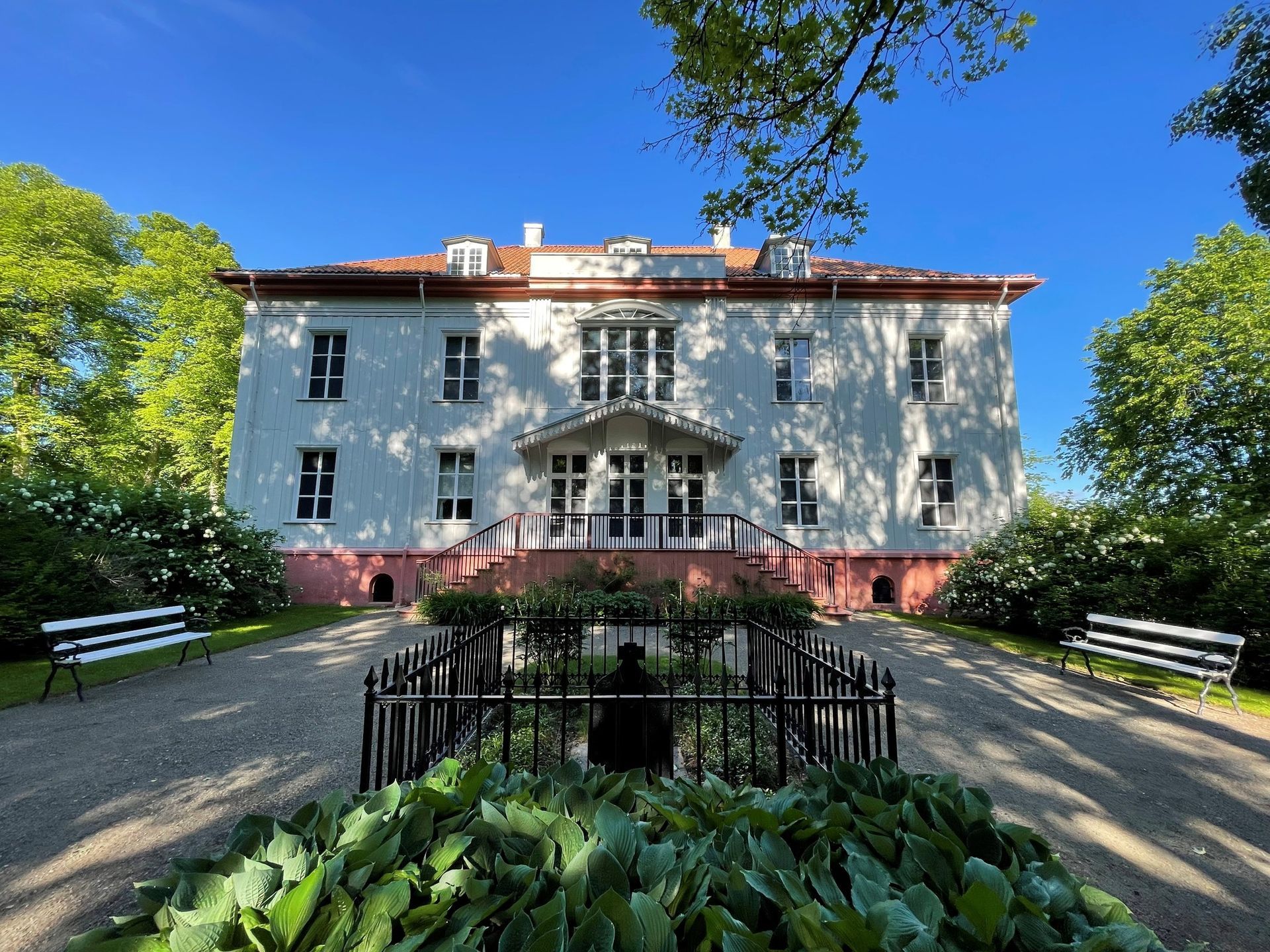 Hvitt herskapshus med rosa detaljer og rødt tak, sett fra en grussti omkranset av busker.