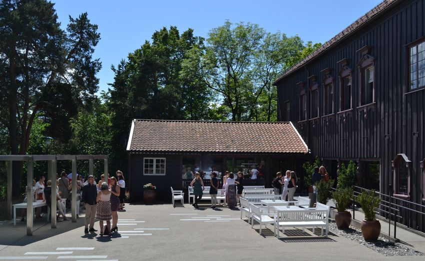 Et mørkebrunt hus med en utsmykket døråpning ligger bak et fargerikt hagebed med lilla og hvite blomster under en blå himmel.