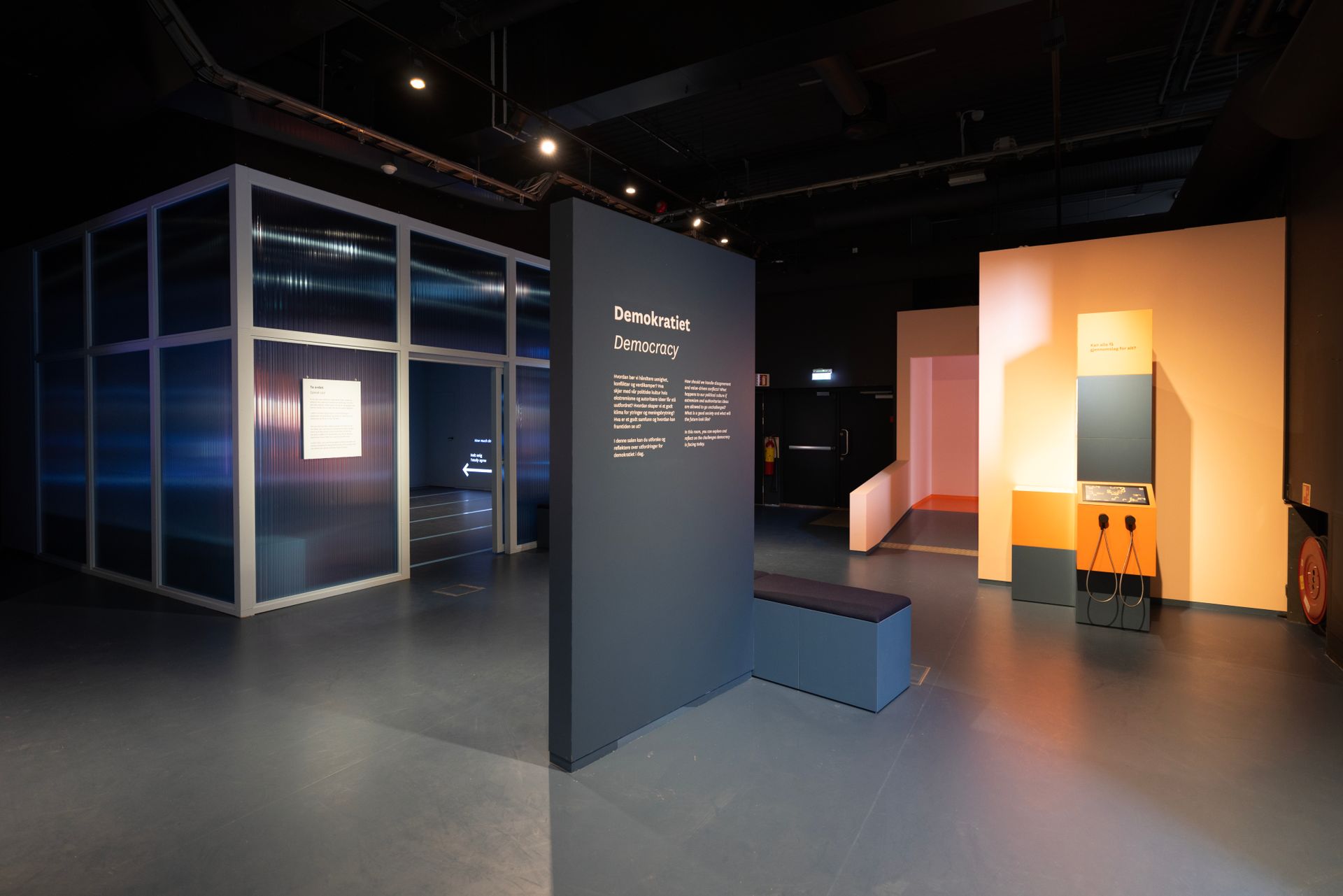 Interiør i en utstilling med en mørkegrå vegg, glassstruktur og oransje og grå strukturer.