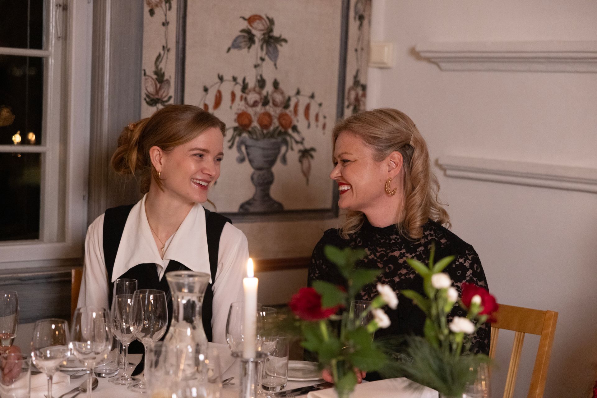 To kvinner smiler til hverandre ved et middagsbord opplyst av levende lys. Blomsterdekorasjoner henger på veggen.